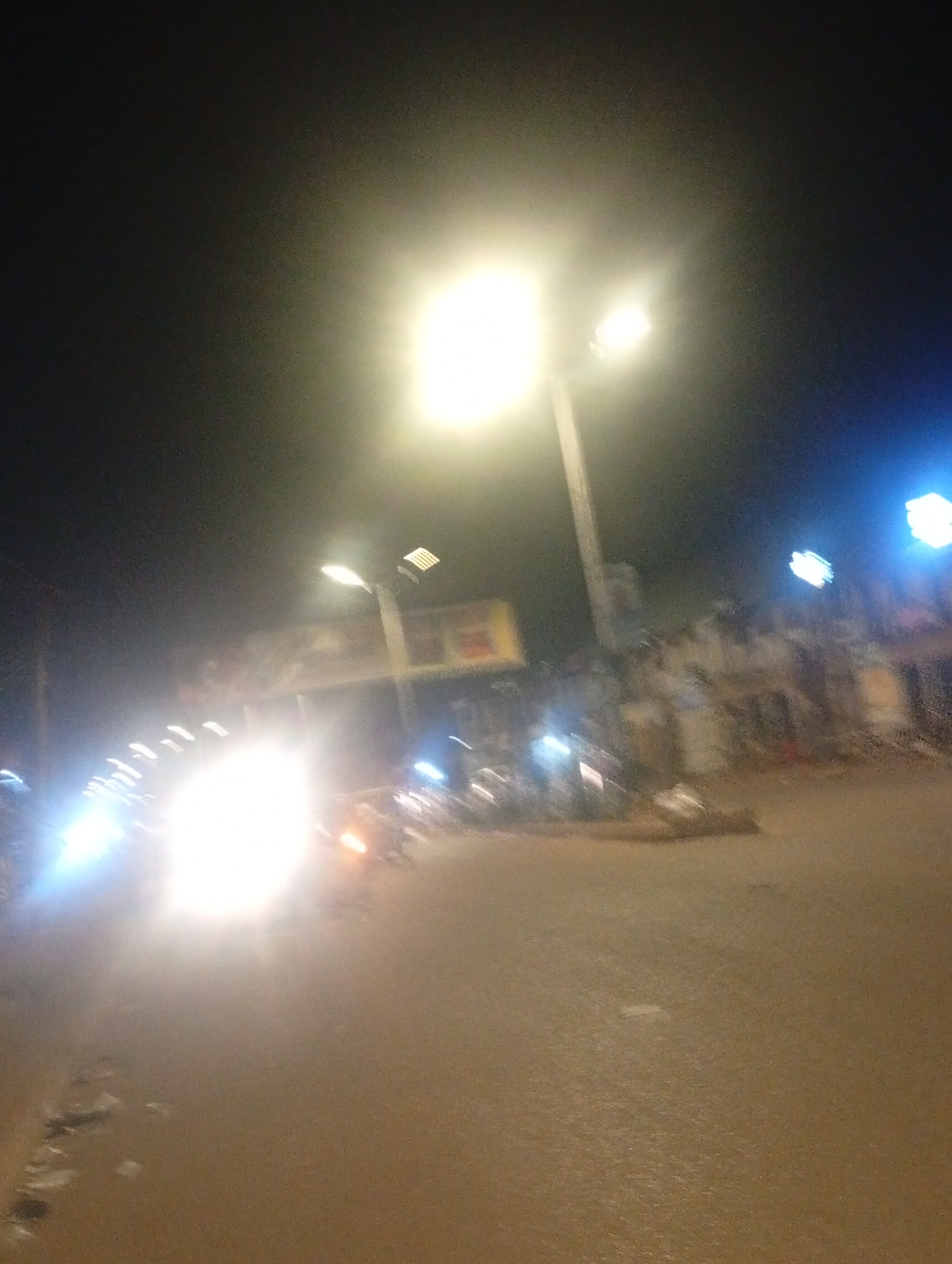 The image depicts a street scene at night in Bukuru, Nigeria. The scene is illuminated by bright streetlights and the headlights of vehicles. The road is paved and appears wet, possibly from rain, reflecting the lights. In the background, there are buildings and various lights, including what appear to be shop signs and more streetlights, some with a blue hue. Several vehicles, including motorcycles, are visible on the road, suggesting traffic. The overall impression is that of a busy urban street at night.