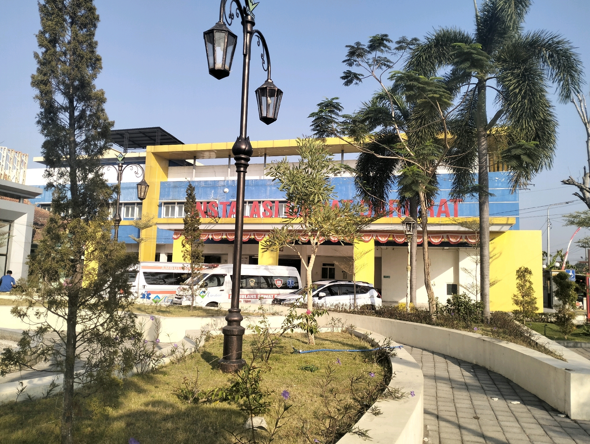 The image captures the exterior of the Bangunsari, Indonesia Public Health Center (Puskesmas). The building features a prominent yellow facade with blue and white accents. Two ambulances are parked in front of the building. A person in a blue shirt is sitting on a concrete ledge on the left side of the image, and another person is visible near the right edge of the frame. The foreground shows a landscaped area with grass, small trees, and a decorative lamp post. A paved walkway curves through the scene. The sky is clear and blue, indicating a daytime setting.