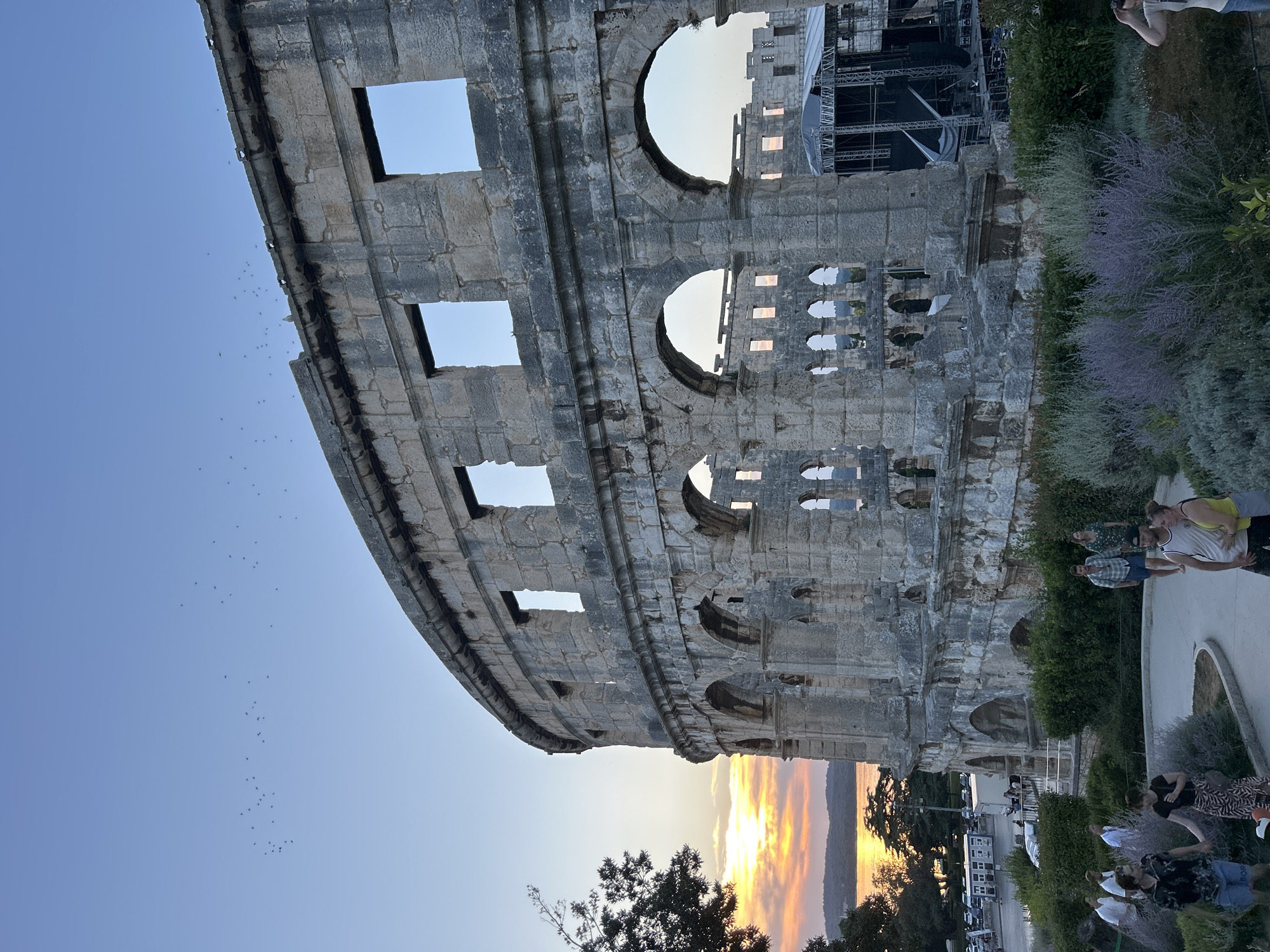 The image depicts a large, ancient stone amphitheater in Pula, Croatia, during sunset. The amphitheater, made of weathered stone blocks, features multiple levels of arches and rectangular openings. A stage setup with scaffolding and lighting equipment is visible within one section of the amphitheater. The sky is a clear blue with a hint of orange and yellow from the setting sun visible over a body of water in the background, reflecting the warm colors. A line of trees borders the water. In the foreground, a paved walkway winds through lush green and purple shrubbery. Several people are walking on the path and around the amphitheater's perimeter, dressed in casual summer clothing. Some are walking in pairs, while others are in small groups. The overall atmosphere is serene and picturesque, capturing the historical grandeur of the amphitheater against a beautiful natural backdrop.