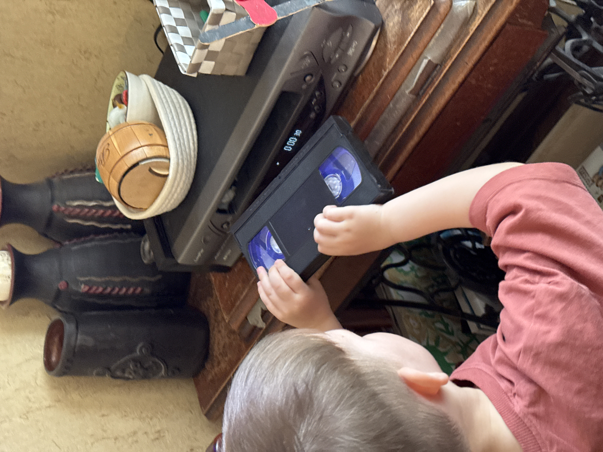 A child is interacting with a VCR player on a wooden surface. The child is holding a VHS tape in their hands, appearing to attempt to insert it into the player. A decorative wooden barrel is next to the player. The location of the image cannot be definitively determined to be in Moscow, Russia, as there are no location-specific visual cues.