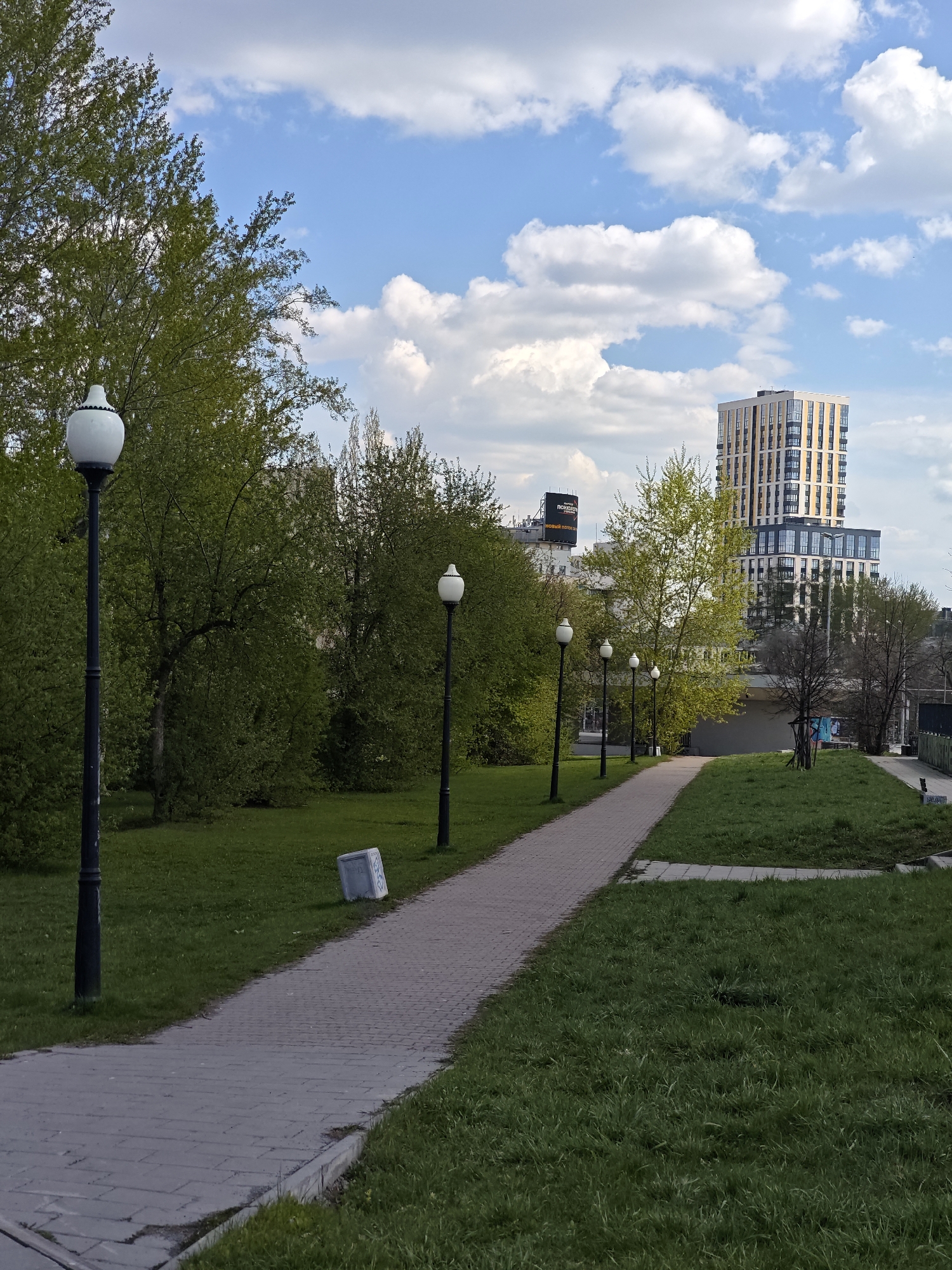 The image depicts a park scene in Yekaterinburg, Russia. A paved pathway lined with lampposts runs through a grassy area with trees. In the background, a tall modern building with a beige and light-colored facade stands out against a blue sky dotted with white clouds. A gray stone block sits near the pathway. The environment suggests a typical urban park setting. No people are visible.