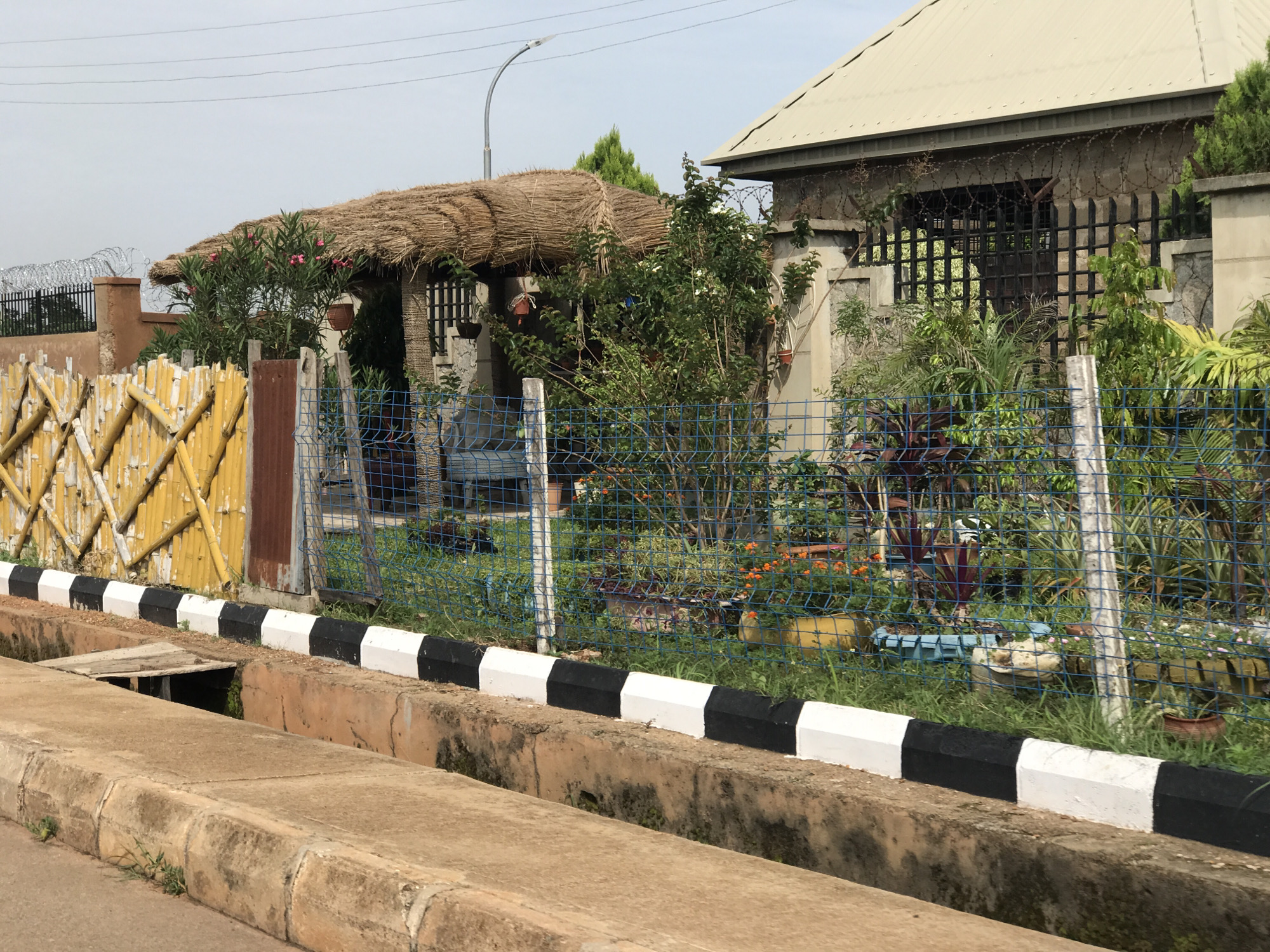 The image depicts a street scene in Jos, Nigeria. In the foreground, a concrete drainage channel runs parallel to the road. The channel has a raised, weathered concrete edge. Along the edge of the road, a curb is painted with alternating black and white stripes. Beyond the curb, a blue wire mesh fence encloses a property.
Behind the fence, a thatched-roof structure, possibly a gazebo or an open-sided shelter, is visible. This structure is adorned with hanging potted plants. A lush garden with various green plants and small orange flowers can be seen within the fenced area. To the right, a building with a beige, sloped roof is partially visible. The building appears to be constructed with concrete blocks and has a decorative metal grille on its upper section, topped with coiled barbed wire.
To the left of the thatched structure, a fence made of yellow-painted bamboo-like poles is present. The overall environment suggests a residential or commercial area with a mix of natural elements and security features. The sky is overcast, indicating a cloudy day.