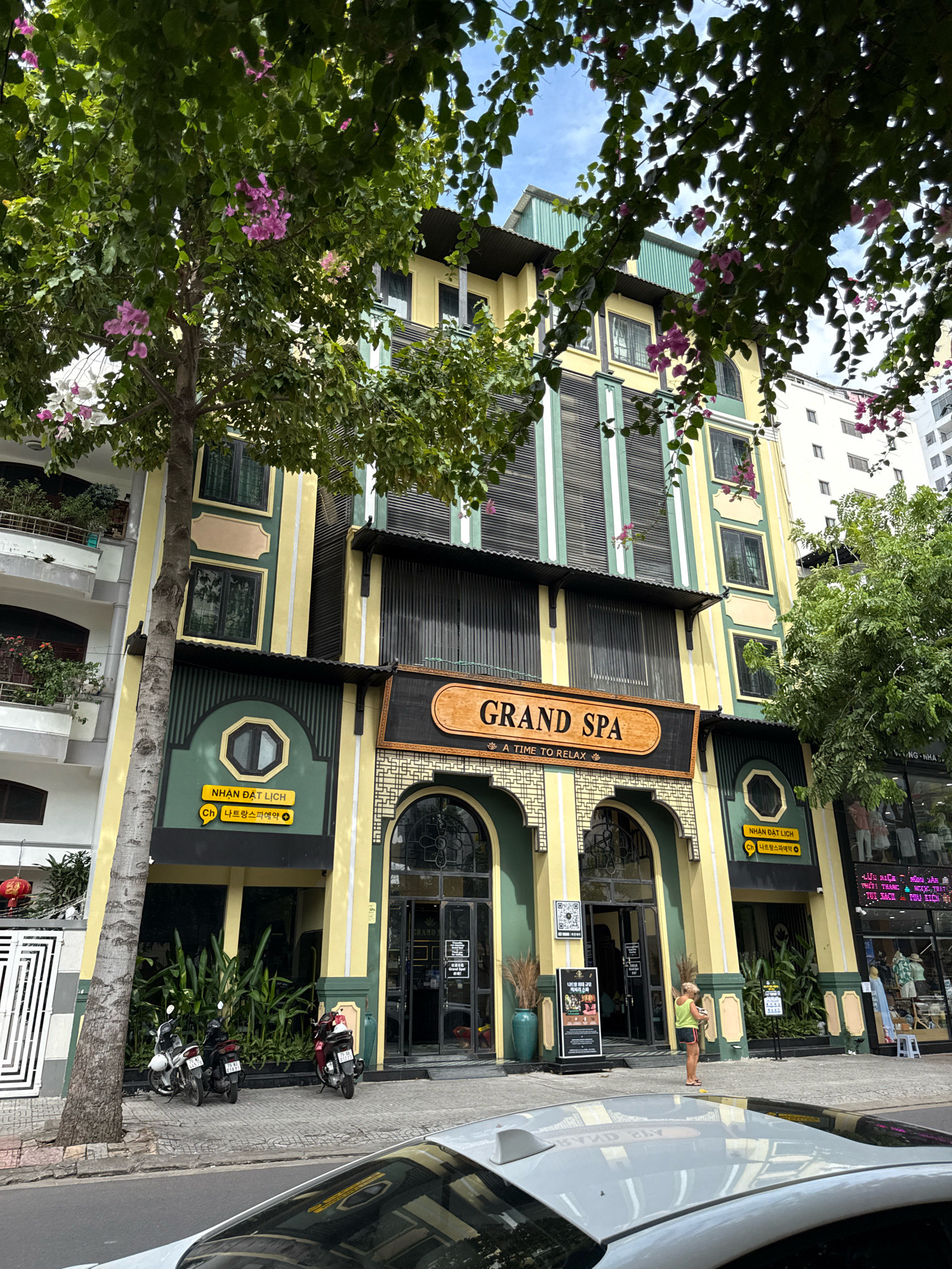 The image shows the exterior of a building labeled "GRAND SPA" with the tagline "A TIME TO RELAX" in Nha Trang, Vietnam. The building is multi-story with a yellow and green facade, featuring large arched entrances. A tree with pink flowers partially obscures the upper floors. In front of the spa, there is a sidewalk with several parked motorcycles. A woman in a yellow tank top and shorts is standing on the sidewalk near the entrance, looking at a sign. Adjacent to the spa are other buildings, including a white high-rise. The overall scene depicts a street view of a commercial area in the city.
