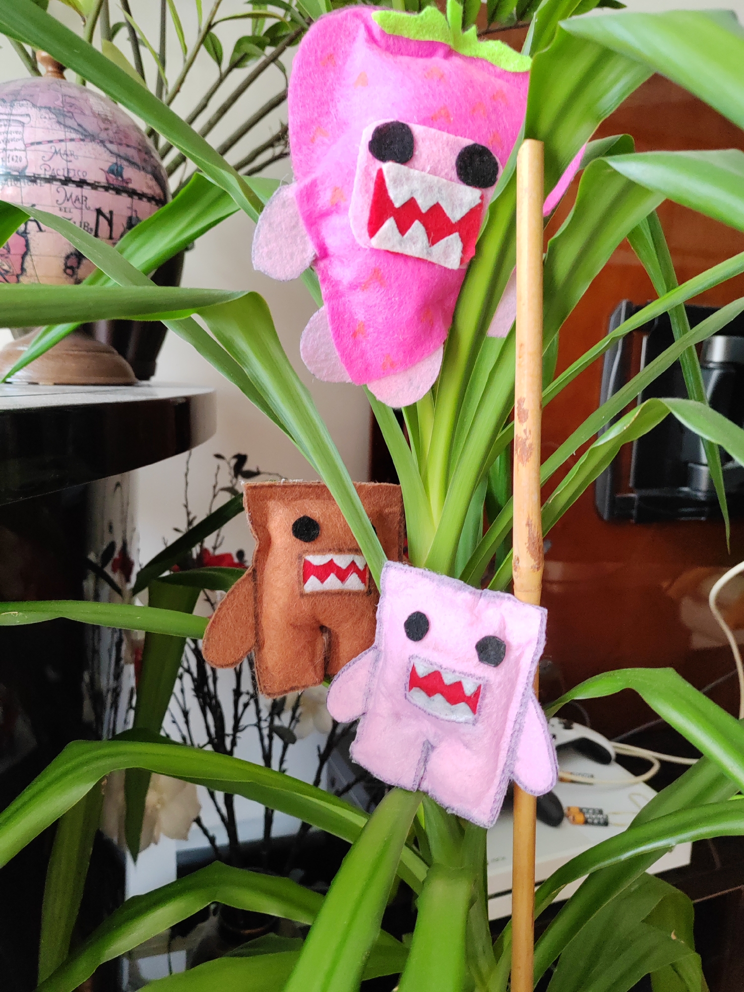 Three handmade felt figures are suspended among the green leaves of a houseplant. The topmost figure is a pink strawberry with a smiling mouth and black eyes. Below it, to the left, is a brown, square-shaped figure with a mouth and black eyes. To the right and slightly lower is a pink, square-shaped figure, similar in design to the brown one, also with a mouth and black eyes. A thin wooden stick passes vertically through the leaves and figures, providing support. In the background, a pink globe is partially visible on the left, and electronic devices and wires are on the right. The scene appears to be indoors, possibly in a domestic setting. The image does not provide any specific information to locate it in Kemerovo, Russia.
