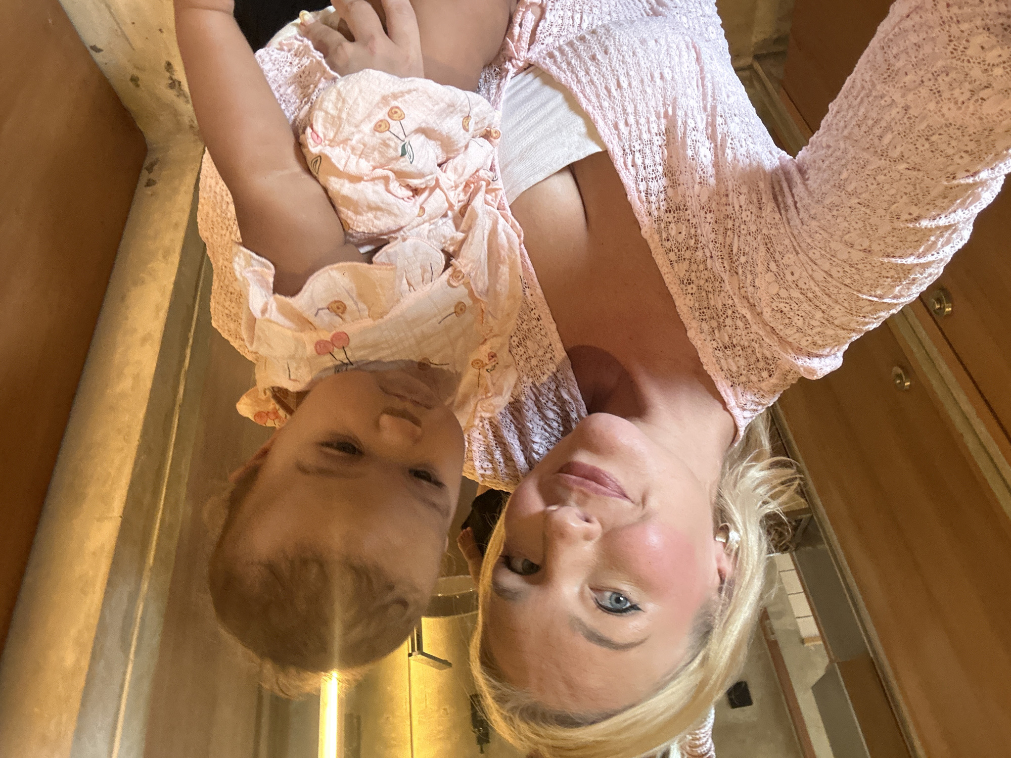 A blonde woman with blue eyes and full lips, wearing a pink lace cardigan and a white tank top, is holding a baby in a pink cherry-patterned onesie. The woman is looking at the camera with a slight smile, while the baby is looking down with a serious expression. They are standing in what appears to be a room with wooden doors and warm lighting, possibly in North Kuta, Indonesia. The background is blurred, but suggests an indoor setting with some architectural features.