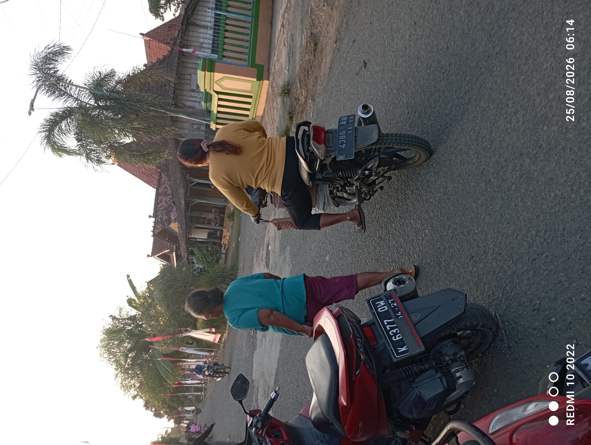 This is a photograph taken in Dukuhseti, Indonesia, on August 25, 2026, at 06:14. The image captures a street scene at dusk, with two women on motorcycles and several other individuals in the background.
In the foreground, a red motorcycle is parked on the left side of the road. To its right, a woman wearing a teal t-shirt and maroon shorts stands facing away from the camera. Further down the road, a second woman, dressed in a yellow long-sleeved shirt and black pants, rides a motorcycle. Her hair is pulled back in a ponytail.
The street is lined with buildings, some of which have traditional Indonesian architecture with thatched roofs. Flags are visible along the side of the road, suggesting a possible local celebration or event. Trees, including palm trees, are also present, contributing to the tropical atmosphere. The road surface appears to be asphalt. The lighting suggests early morning or late evening. Other people and motorcycles are visible in the distance, indicating a populated area. The overall scene depicts a typical day in a village or small town in Indonesia.