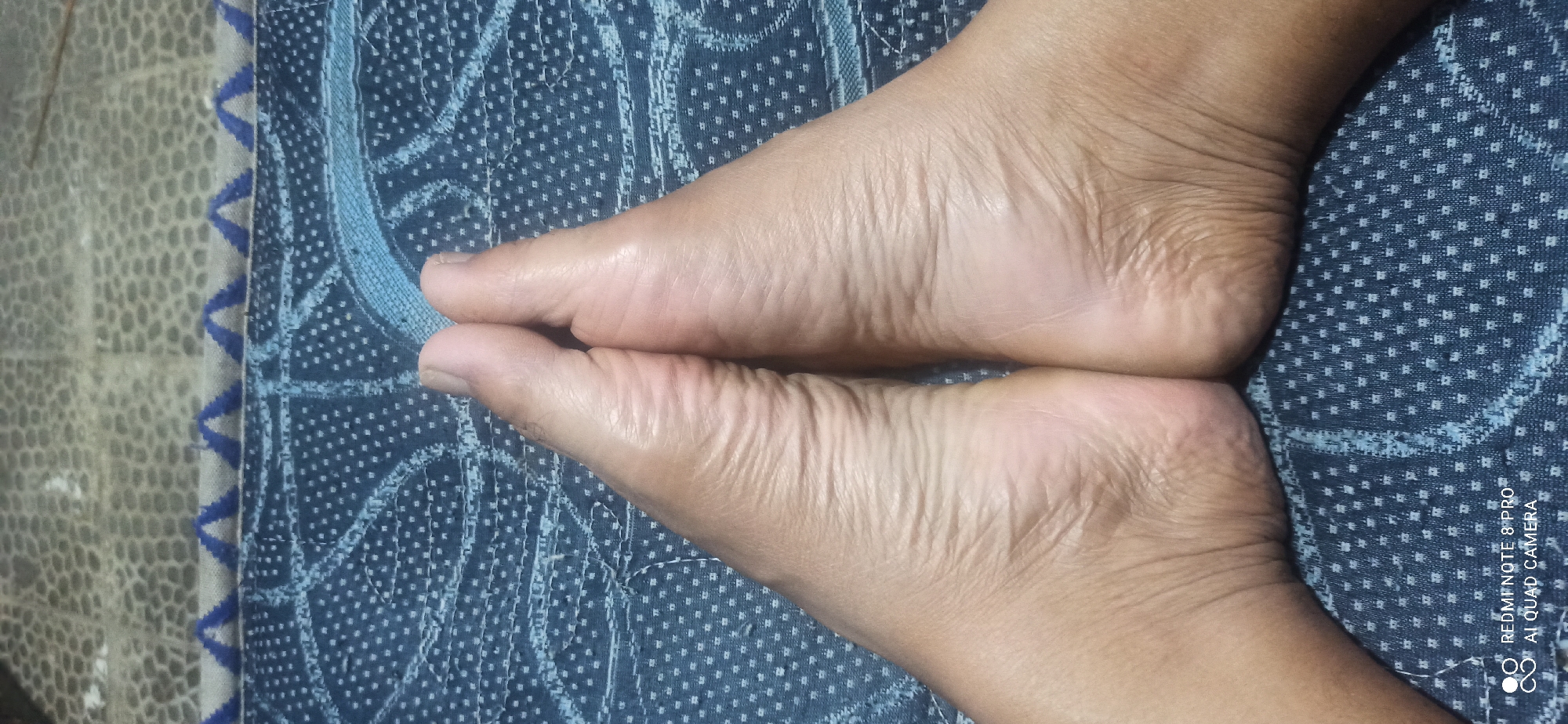A close-up, overhead view shows the soles of two feet pressed together, forming a V-shape. The skin on the feet is wrinkled and tanned. The feet are positioned on a dark blue fabric with a repeating white dotted pattern and some lighter blue abstract designs. In the background, a greyish textured surface is visible. The bottom left corner of the image contains a watermark that reads "REDMI NOTE 8 PRO AI QUAD CAMERA". The image was taken in Sedati Agung, Indonesia.