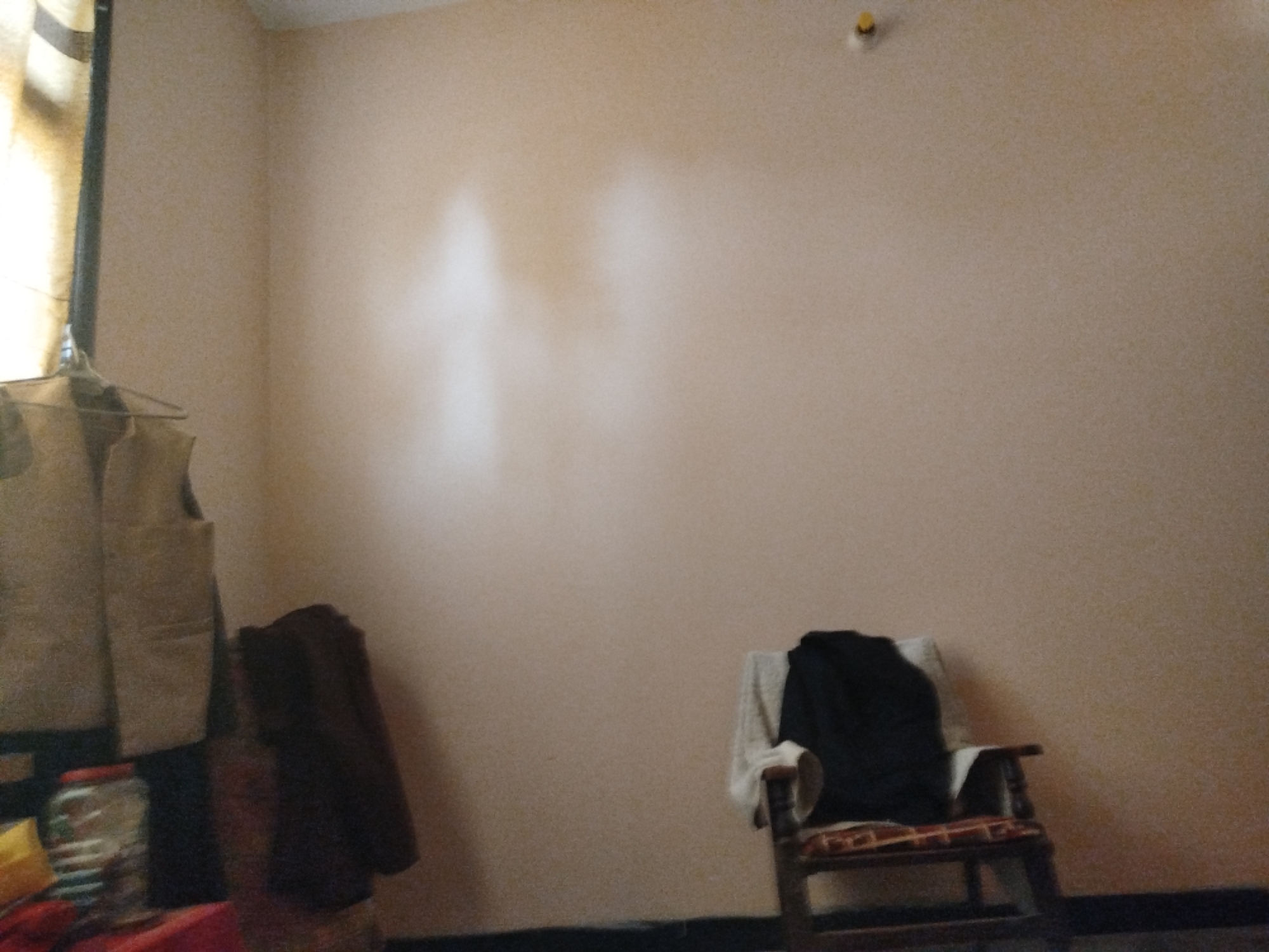 The image depicts an indoor scene within what appears to be a room in Lassan, Pakistan. The walls are painted a pale, peachy color, with some subtle variations in tone suggesting texture or perhaps lighting effects. In the upper right corner, a small yellow circular object is affixed to the wall, possibly a light fixture or a decorative element.
On the left side of the frame, a clothing rack is visible, with a beige vest or jacket hanging from it. Next to the clothing rack, a dark brown garment is draped over an object, and a clear glass jar with a red lid is present on a surface.
In the lower right portion of the image, a wooden chair with a patterned cushion is situated. A dark-colored garment, likely a sweater or jacket, is placed over the back of the chair.
The floor appears to be dark. The overall impression is of a casual, lived-in space. The lighting is diffused, casting a soft glow on the walls and objects. No individuals are present in the frame.
