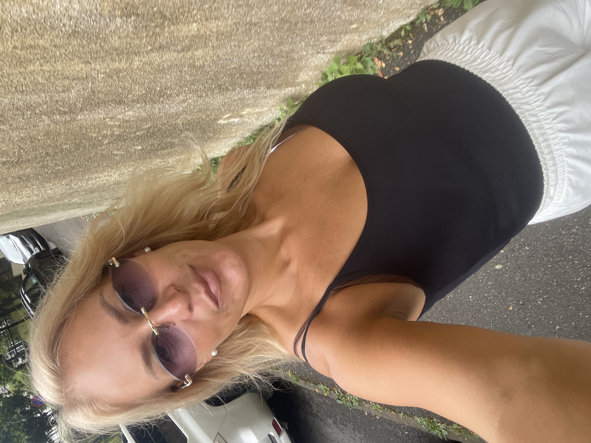 A woman with blonde hair and sunglasses is taking a selfie outdoors. She is wearing a black tank top and white pants. Behind her, a white car is parked on the side of the road, and a concrete wall is visible. The setting appears to be a street in Sochi, Russia.