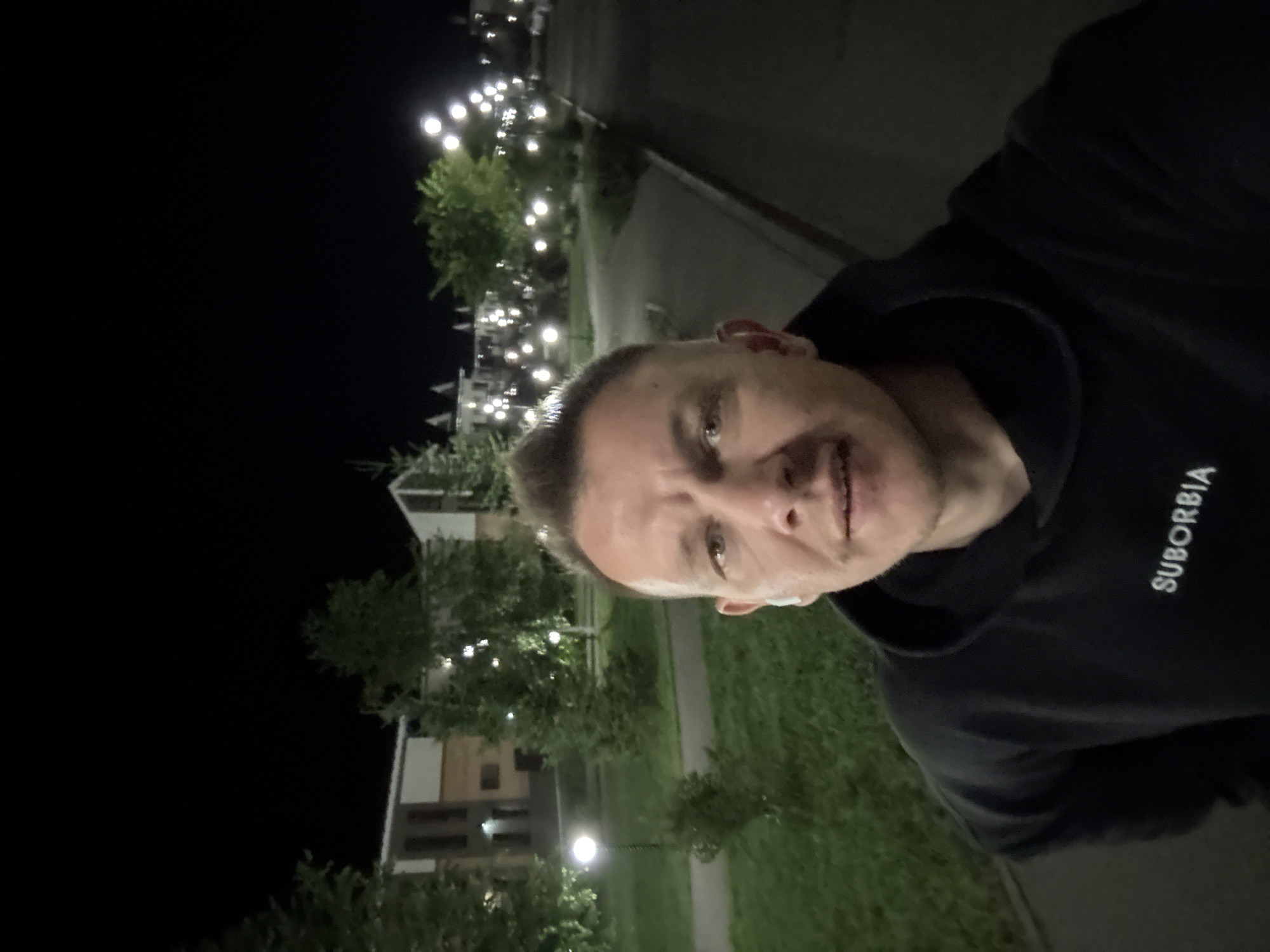 A man, with short blonde hair, is taking a selfie at night on a street in Markova, Russia. He is wearing a black hoodie with "SUBURBIA" written on the chest. The background shows a residential area with modern-looking buildings, trees, and streetlights. The lighting is dim, indicating it is nighttime. The man is looking directly at the camera. The image appears to be taken outdoors, likely on a sidewalk or pathway.