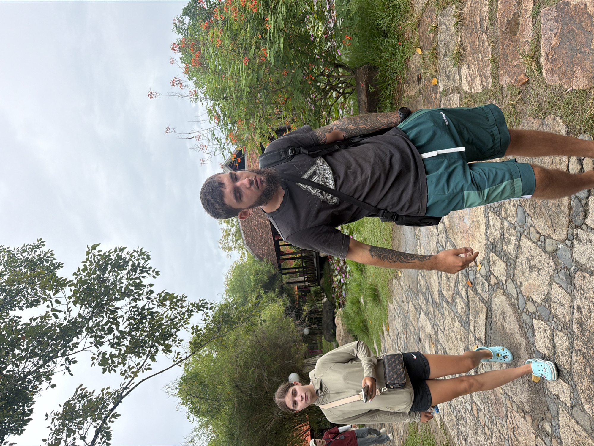A young woman with dark hair stands on a cobblestone path, holding a smartphone and looking forward. She wears a light-colored hooded jacket, black shorts, and light blue Crocs. To her right, a man with a beard and tattoos walks along the path, wearing a dark t-shirt with an Adidas logo and green shorts. He carries a black backpack. In the background, trees, flowering bushes with orange blooms, and a traditional Vietnamese building with a tiled roof are visible. The ground is a mixture of cobblestones and grass. The scene appears to be in a park or garden setting in Nha Trang, Vietnam.