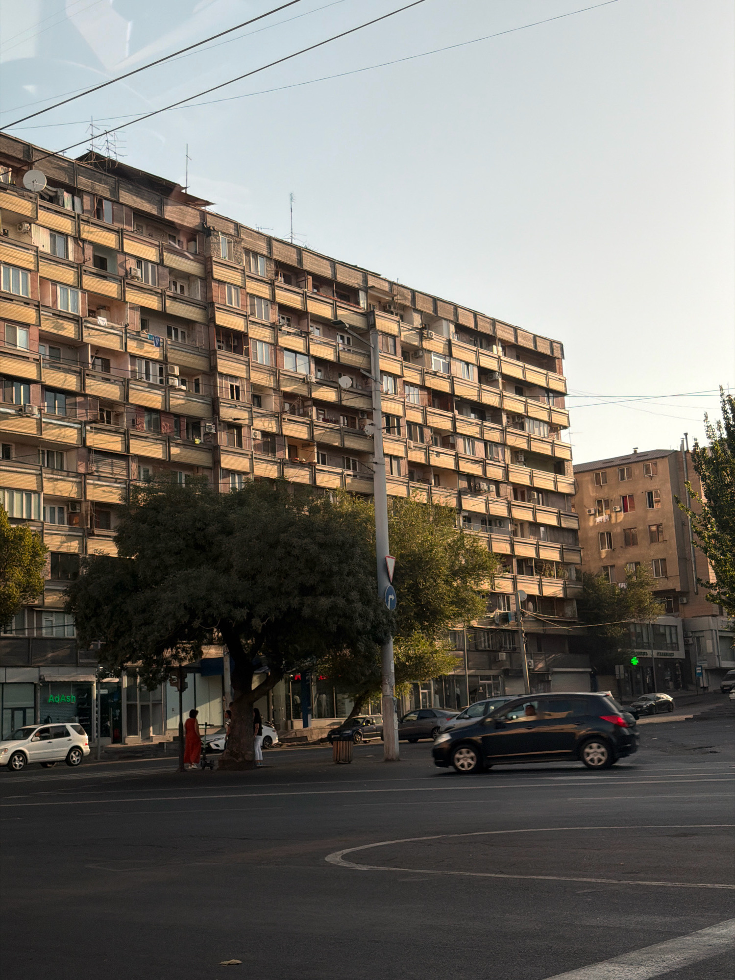 The image depicts a street scene in Yerevan, Armenia, likely in the evening given the warm, low sunlight. Dominating the left side of the frame is a large, Soviet-era apartment building with many balconies and windows, constructed with a beige facade and dark-colored trim. In front of this building, a large, leafy tree provides shade. To the right of the apartment building, a smaller, slightly darker building with similar window arrangements is visible.
On the street, several cars are present. A white SUV is parked on the left, and a dark hatchback is driving towards the right of the frame in the foreground. In the middle ground, a person in an orange garment stands near the tree, with other figures and a white car visible behind them. The street itself is paved, with lane markings indicating traffic flow. Power lines are strung across the sky, a common feature in urban environments. The sky is clear and a pale blue, transitioning to a warmer hue near the horizon. The overall impression is one of an active, urban environment.