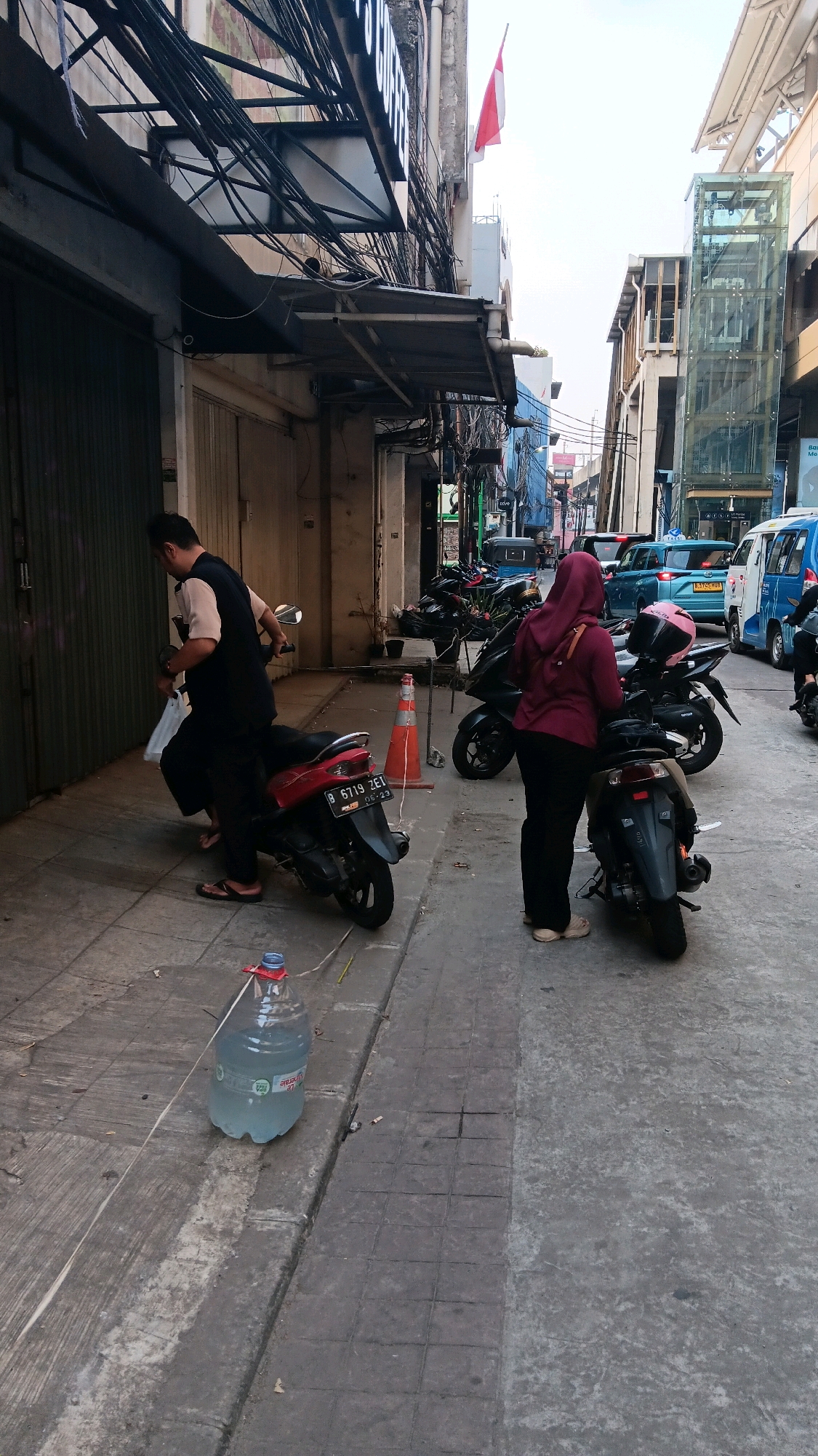 The image shows a street scene in South Jakarta, Indonesia, with several motorcycles parked along the sidewalk. A man in a black vest over a tan shirt is standing next to a red motorcycle. He appears to be holding a white plastic bag. Further down the street, a woman wearing a maroon hijab and black pants is standing next to a black motorcycle. A large plastic water jug is on the sidewalk in the foreground. Buildings line the street, with overhead electrical wires creating a dense network above. In the background, there are cars, a blue bus, and an elevated structure that appears to be part of a transportation system. An Indonesian flag is visible flying from a pole on one of the buildings. The overall impression is of a busy urban environment with pedestrian and vehicular traffic.