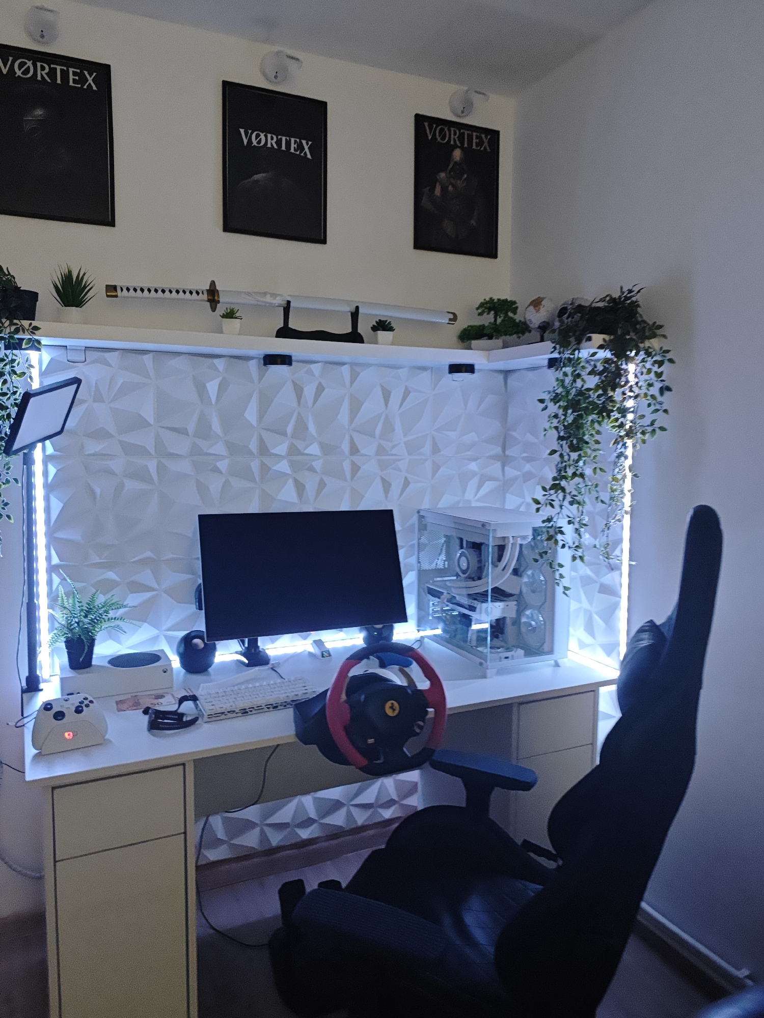 The image shows a gaming setup in a room. The setup includes a computer desk with a white desktop and drawers, a monitor, a white gaming PC case with visible components, a keyboard, a gaming wheel, and a white Xbox controller. Behind the desk is a wall with a 3D geometric pattern. LED strip lights are positioned along the edges of the wall. Above the desk are three framed posters with the word "VORTEX" and a shelf with decorative plants and a katana. A black gaming chair is positioned in front of the desk. The location, Stambolovo, Bulgaria, is not evident from the image.