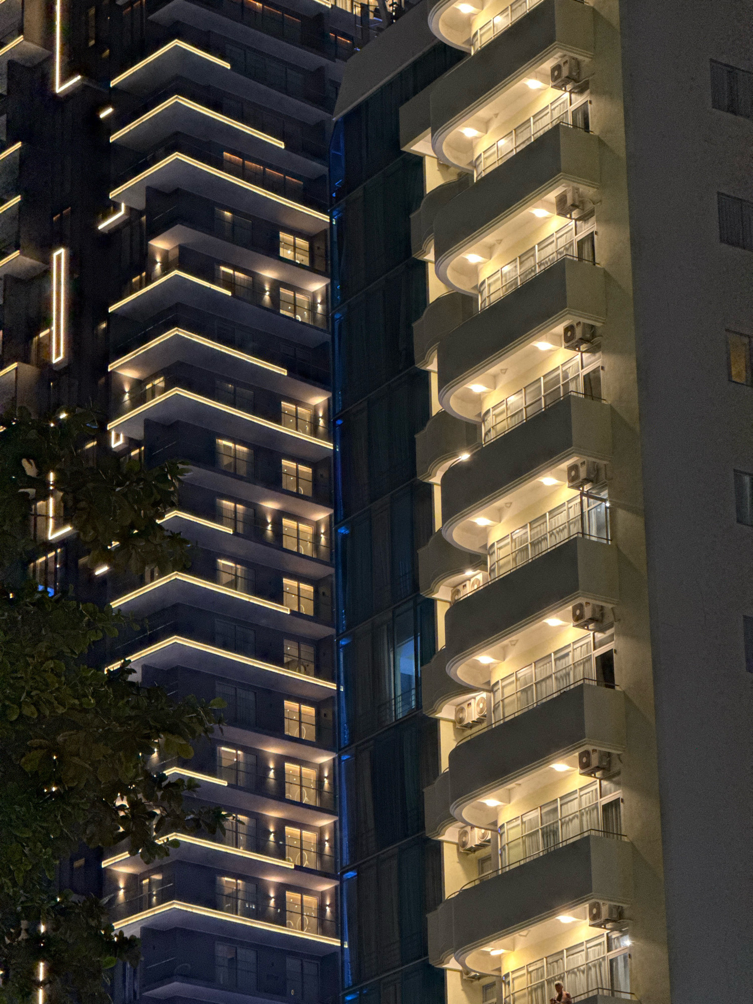 The image depicts a nighttime view of two adjacent high-rise buildings in Nha Trang, Vietnam. The building on the left is modern with dark facade and illuminated by linear LED lights tracing the edges of balconies and vertical accents. The building on the right features lighter colored balconies with curved edges, each illuminated by multiple lights from above. Multiple air conditioning units are visible on the balconies. A portion of a leafy tree is in the foreground on the left, partially obscuring the lower levels of the left building. The windows of both buildings show interior lights, suggesting occupancy.