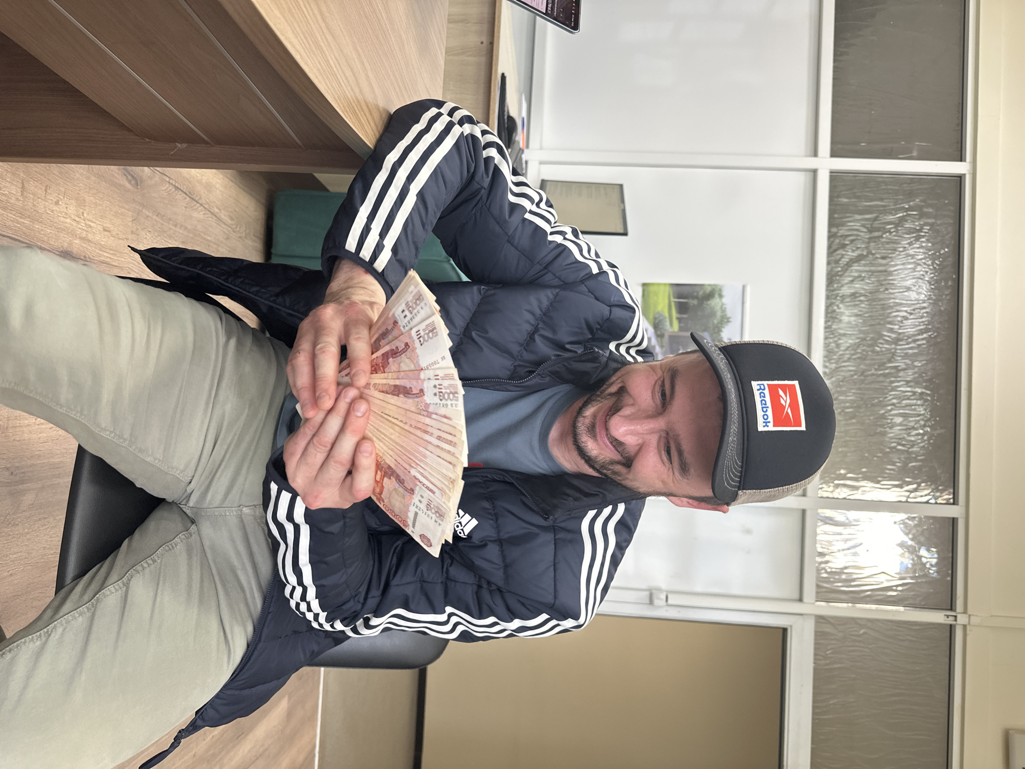 A man is sitting on a black chair in a room. He's wearing a dark blue jacket with white stripes, a gray t-shirt, and light khaki pants. He has a black and tan baseball cap with a red and white Reebok logo. He's holding a large fan of reddish-orange banknotes, which appear to be Russian rubles, likely 5000 ruble notes. The man is smiling. Behind him is a desk, a window with a framed picture on the sill, and an office-style window structure. The location is not specified as Izhevsk, Russia.