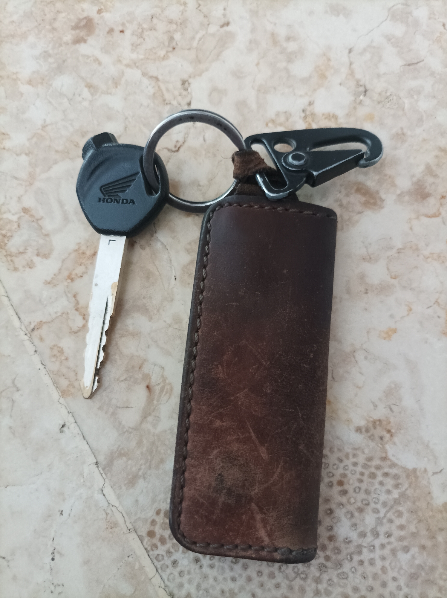 A Honda motorcycle key with a black fob featuring the Honda logo is attached to a silver keyring. Also on the keyring is a black metal carabiner clip and a brown leather key pouch. The objects are lying on a marble-patterned surface with varying shades of beige and white. The scene is photographed from a top-down perspective. There is no indication of the location in Nganjuk, Indonesia.