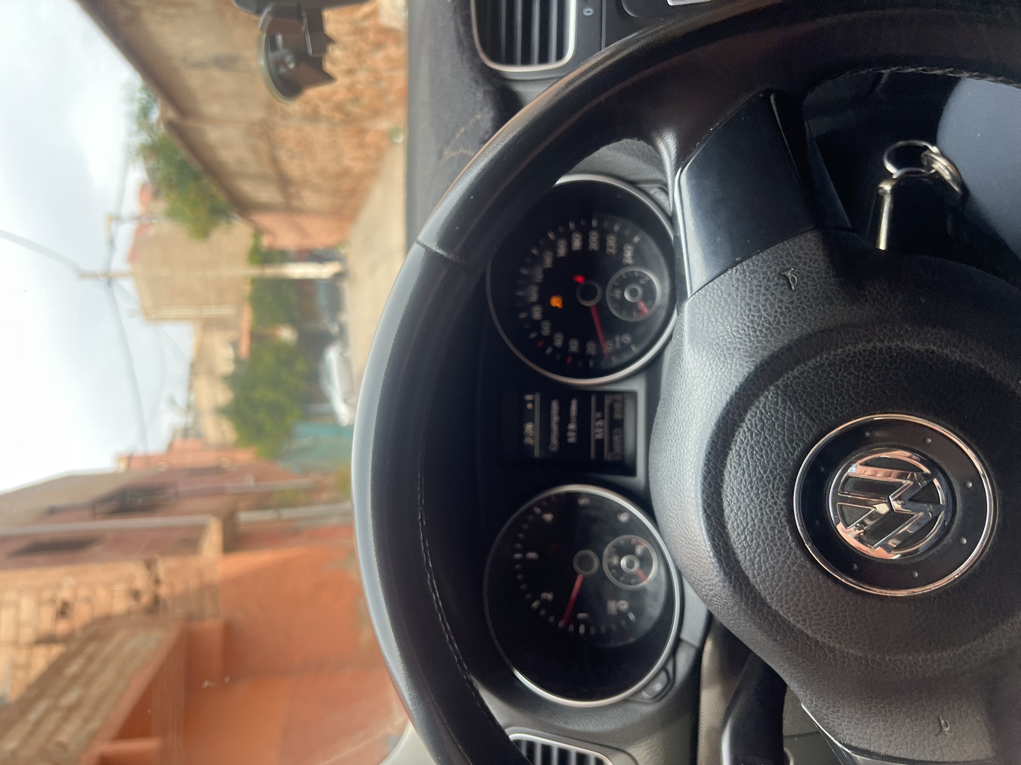 The image shows the interior of a Volkswagen vehicle, specifically the steering wheel and dashboard, with a partial view through the windshield revealing a street scene. The steering wheel is black leather with a prominent Volkswagen logo in the center. Keys are present on the steering column. The dashboard includes the speedometer and other gauges, with indicator lights illuminated. Through the windshield, a blurred street scene is visible, featuring buildings constructed with red brick or clay, characteristic of the city of Ouaouizert, Morocco. The buildings appear to be two or three stories high, with some greenery visible. The sky is overcast. The image suggests the vehicle is stationary.