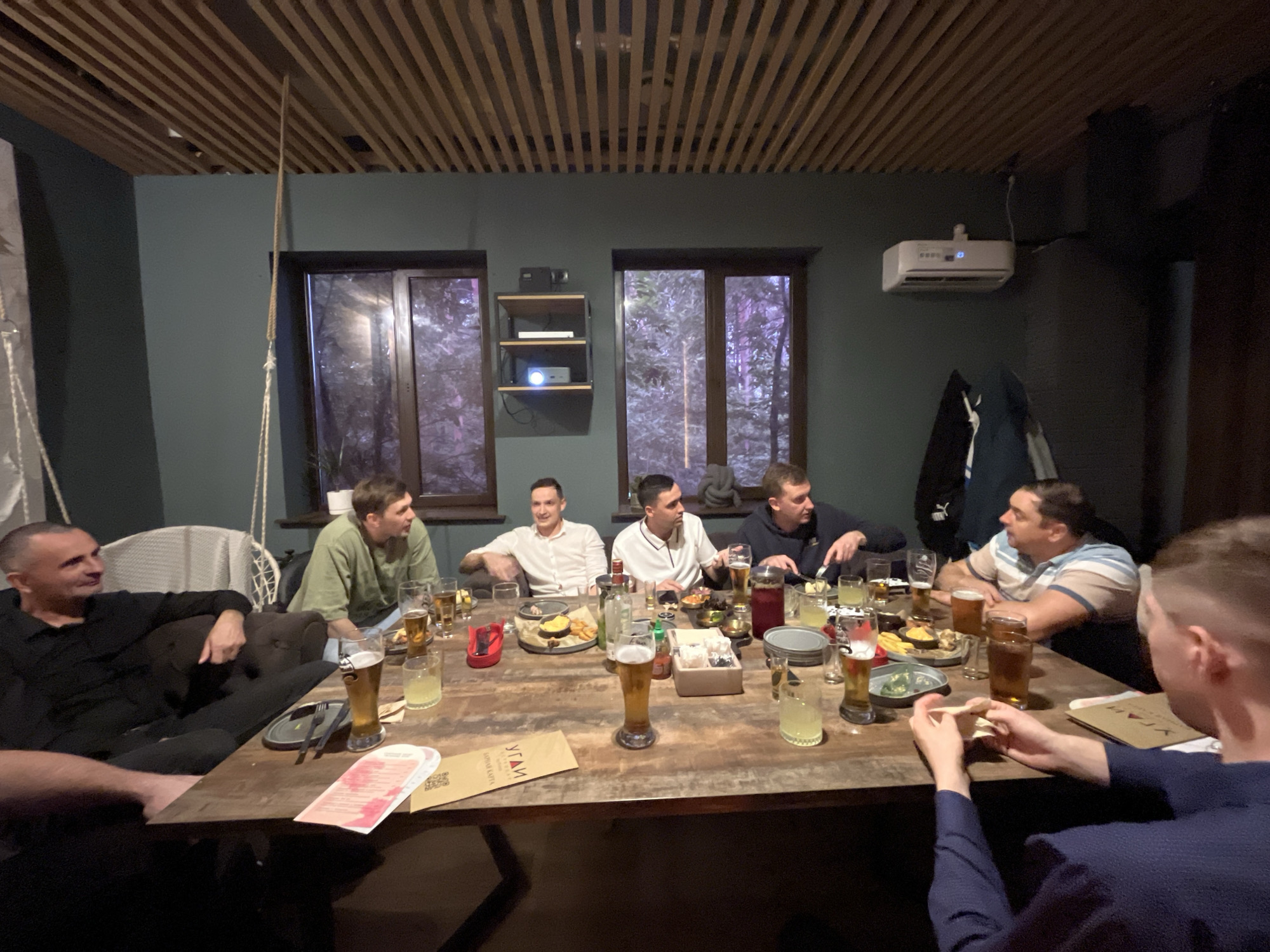 The image depicts an indoor social gathering of approximately nine men seated around a large, rustic wooden table in what appears to be a restaurant or bar in Chaikovsky, Russia. The room features dark green walls, a striking wooden slatted ceiling, and large windows showcasing a dark, possibly wooded, outdoor environment at night.
The men are casually dressed and are engaged in conversation, eating, and drinking. The table is filled with various food platters, including nachos and other appetizers, alongside numerous glasses of beer and other beverages. Bottles, including a clear spirit bottle, and a carafe of dark liquid are also present.
Notable details include a white projector situated on a shelf between two windows, and a white air conditioning unit mounted high on the wall to the right. A rope hangs from the ceiling near a white wicker chair and a dark sofa where one man is reclining. The atmosphere is relaxed, suggesting a casual evening get-together. On a brown menu or brochure on the table, the Cyrillic word "УГЛАИ" (UGLAI) is visible. The scene is illuminated by warm, artificial interior lighting.
