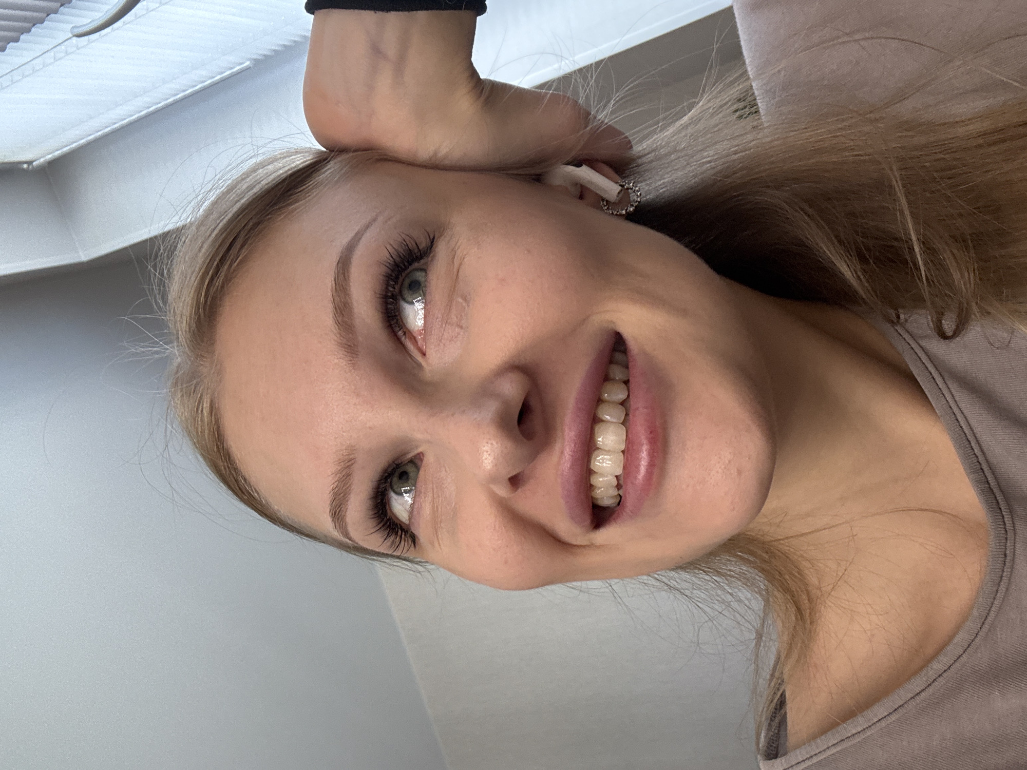 A young woman with blonde hair and blue eyes is captured in a close-up, low-angle selfie. She is wearing a light purple top and has a subtle smile, revealing her teeth. Her right hand is resting on her head, and she is wearing white wireless earbuds and a sparkling hoop earring on her right ear. The background appears to be an indoor setting with a textured wall and a window with blinds. There is no discernible indication of the city of Perm, Russia, in the image.