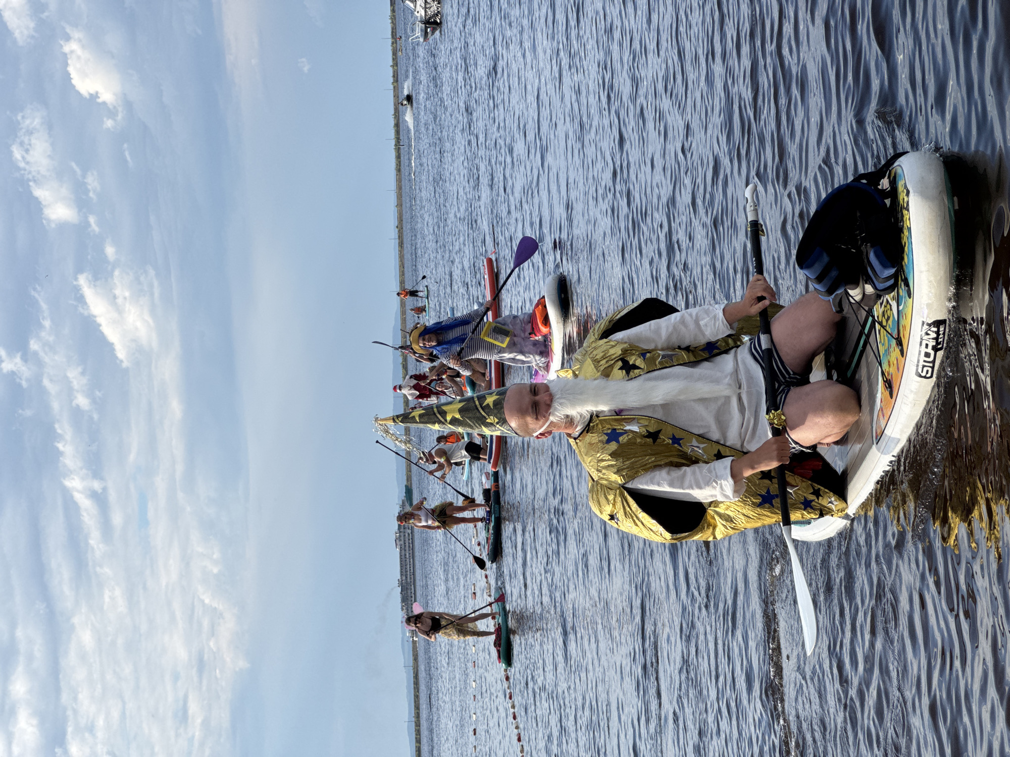 In a wide shot of a body of water, a man dressed as a wizard is seen paddleboarding. He has on a gold robe with stars and a pointy hat. He is holding a paddle. In the background, other people are also paddleboarding, some of whom are in costume. The water is rippled, and the sky is partly cloudy. The scene appears to be taking place during the day. There is no visible text in the image, but the SUP board in the foreground has "STORM LINE" printed on it.