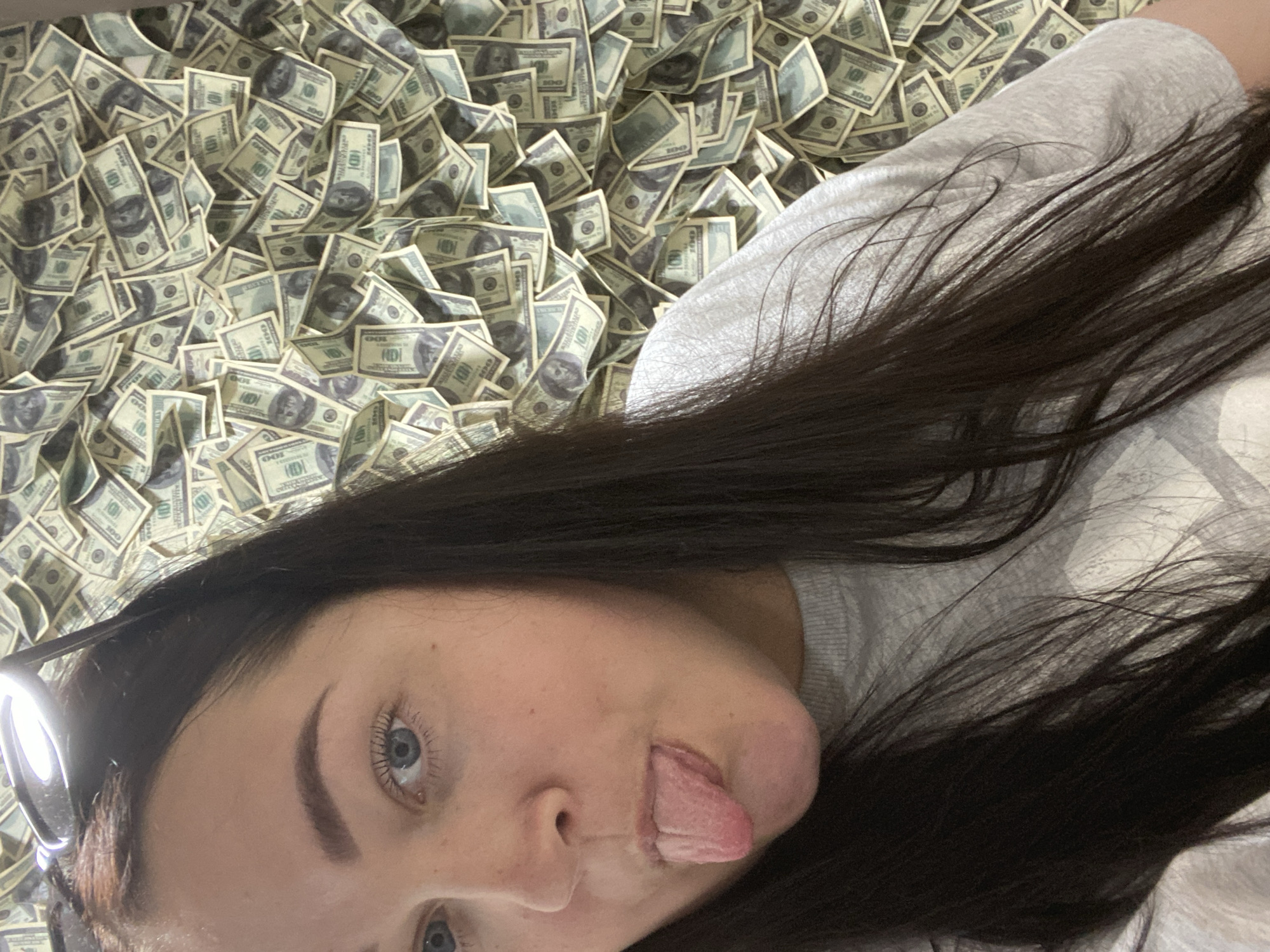 A woman with dark hair is lying on a surface covered in U.S. hundred-dollar bills. She is sticking out her tongue and looking at the camera. The woman is wearing a light gray shirt. Sochi, Russia is not discernable from the available information.
