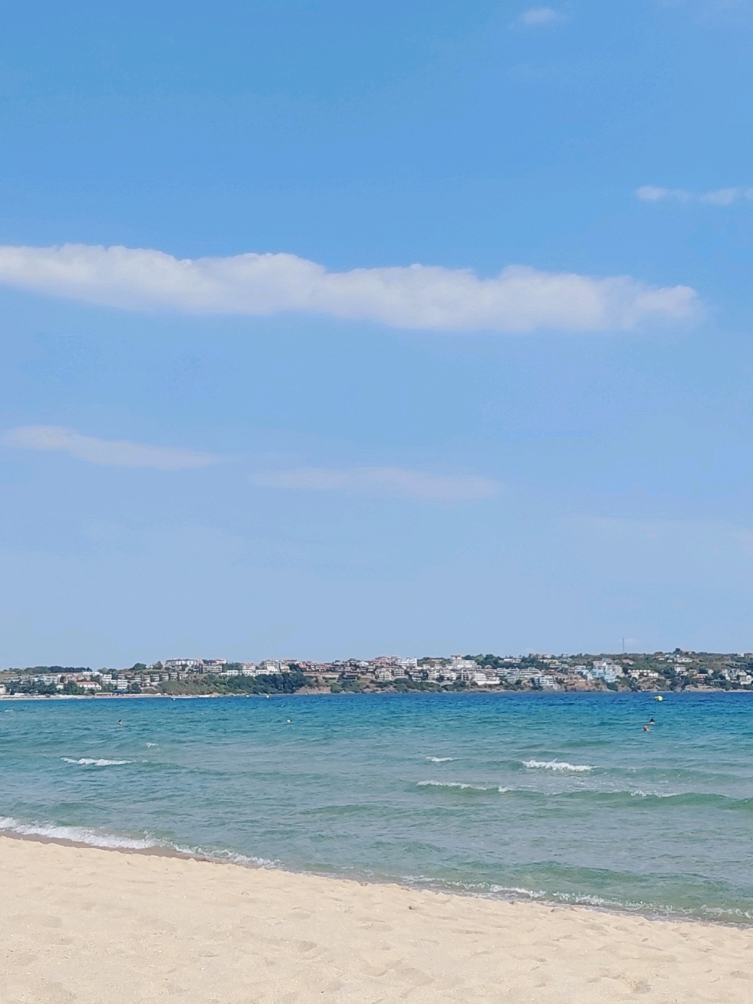 The image depicts a coastal scene in Sozopol, Bulgaria. In the foreground, a sandy beach stretches across the bottom of the frame, meeting the gentle waves of the sea. The water is a clear turquoise, with small white-capped waves rolling towards the shore. In the middle ground, the sea continues, and a distant coastline is visible. Along this coastline, a town or city is situated, with numerous buildings and structures dotting the landscape. The buildings appear to be of varying sizes and colors, with many featuring white facades. Green vegetation covers the hills behind the town, suggesting a natural, hilly terrain. The sky above is a bright blue, with a few scattered white clouds. The overall impression is one of a sunny day at a coastal resort. There are a few indistinct figures in the water, suggesting recreational activity.