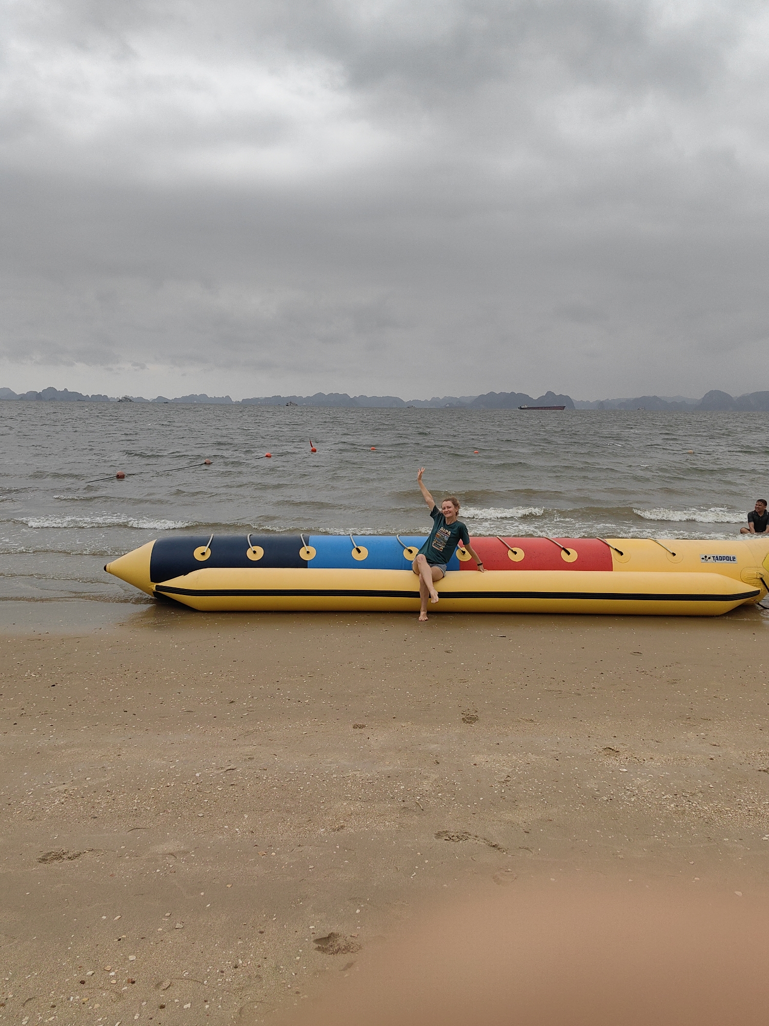 A woman is sitting on a yellow, blue, and red banana boat on a beach in Bai Chay Ward, Vietnam. She is waving her right hand up. There is a dark sky with some islands in the background, and the waves are coming up on the beach. There is another person to the right of the banana boat sitting near the end of the boat.