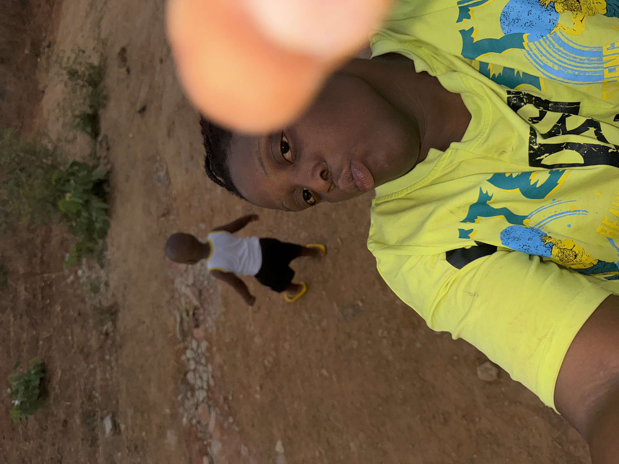 A selfie-style photograph shows a person with dark skin wearing a patterned yellow shirt. Their face is close to the camera with a slightly puckered expression. A blurred finger obscures part of the view.
In the background, a child wearing a white shirt, dark shorts, and yellow sandals is walking on a dirt path. The surrounding environment appears to be a slightly overgrown area with dirt and some sparse vegetation.
The location is not specified. The prompt requested the location in the city of Sabon Tasha, Nigeria, but this cannot be determined from the image itself.