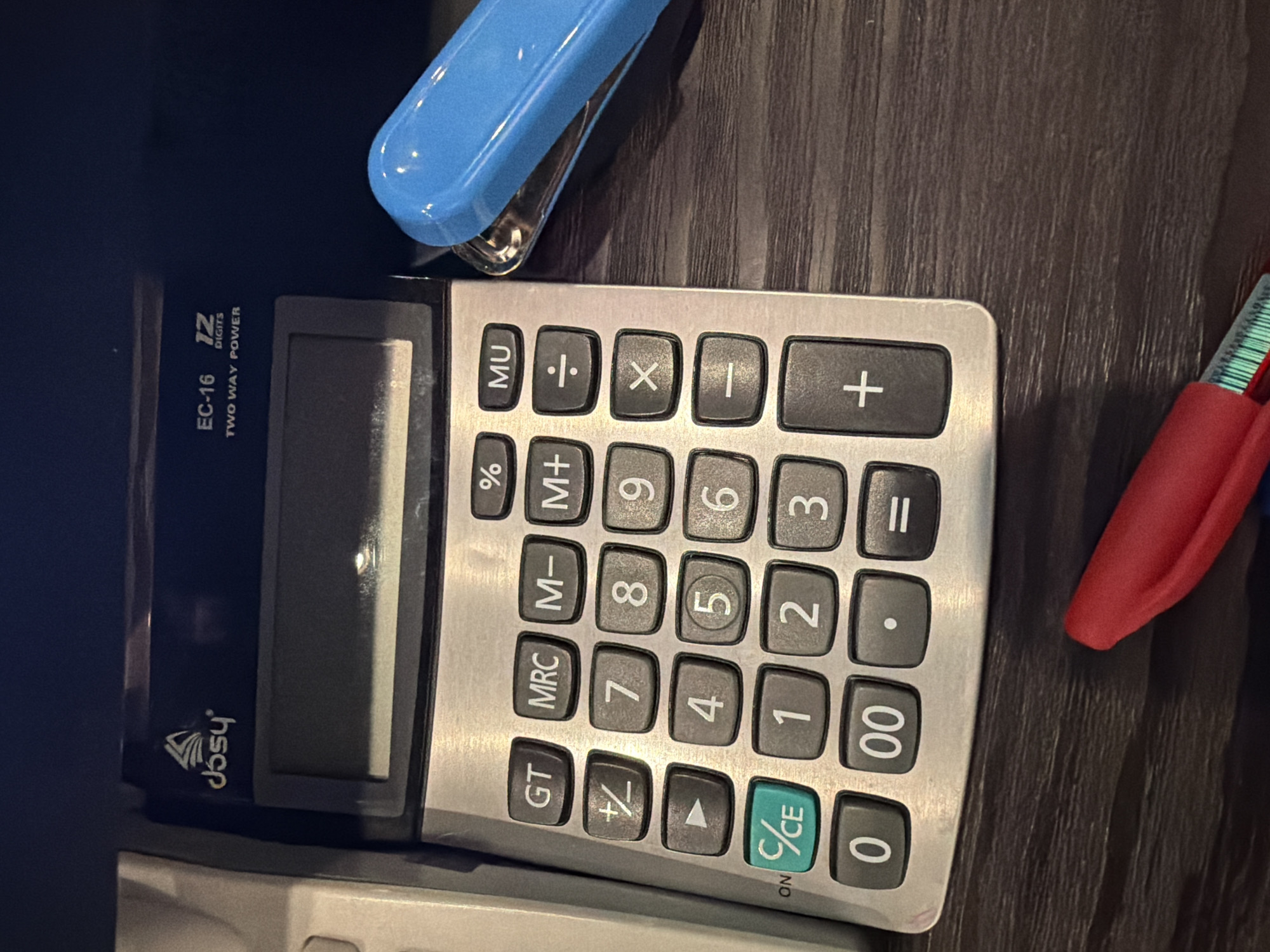 The image shows a close-up, eye-level, overhead view of a desk with office supplies. In the center is a silver calculator with a dark screen and numerous buttons, including numbers 0-9, arithmetic operations, and memory functions. The "C/CE" button is a vibrant green. To the right of the calculator is a bright blue stapler. In the lower foreground, a red pen with green markings is visible. The desk surface is a dark wood grain. The background is dark and out of focus, showing a sliver of what appears to be another office appliance. The scene is indoors and appears to be a workspace. There are no people or animals in the image. The lighting is moderate, highlighting the calculator and stapler. There is no information in the image to suggest a location in the city of Sofia, Bulgaria.