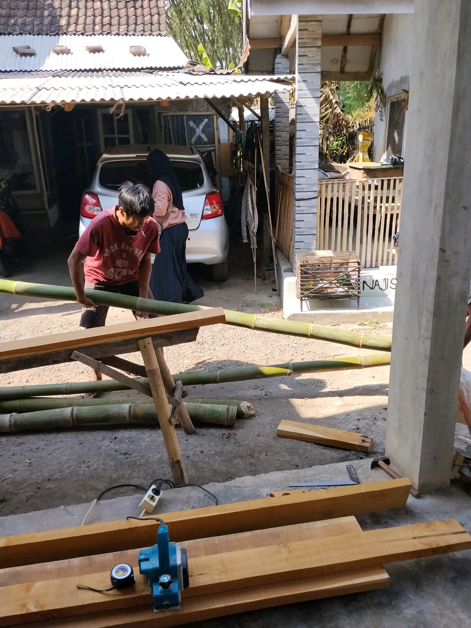 In Panggungan, Indonesia, an outdoor scene unfolds with construction or craft work in progress. A young man, wearing a red t-shirt and dark shorts, is positioned at a sawhorse, holding a long, green bamboo pole. He appears to be working with the bamboo, which is laid out on a makeshift wooden table and on the ground. Nearby, a silver car is parked, partially covered by a corrugated metal awning supported by wooden beams. A woman in a pink hijab and dark skirt stands near the car, facing away from the camera.
The environment is a yard with a concrete floor. Piles of cut lumber are stacked in the foreground, alongside a blue electric planer and a tape measure. Various tools, including a hammer and a saw, are scattered around the lumber. In the background, a wooden fence encloses an area where a birdcage with small birds is visible. The structures suggest a residential setting, with parts of a house visible, including tiled roofing and walls. The overall impression is one of a busy, functional outdoor workspace under the bright Indonesian sun.