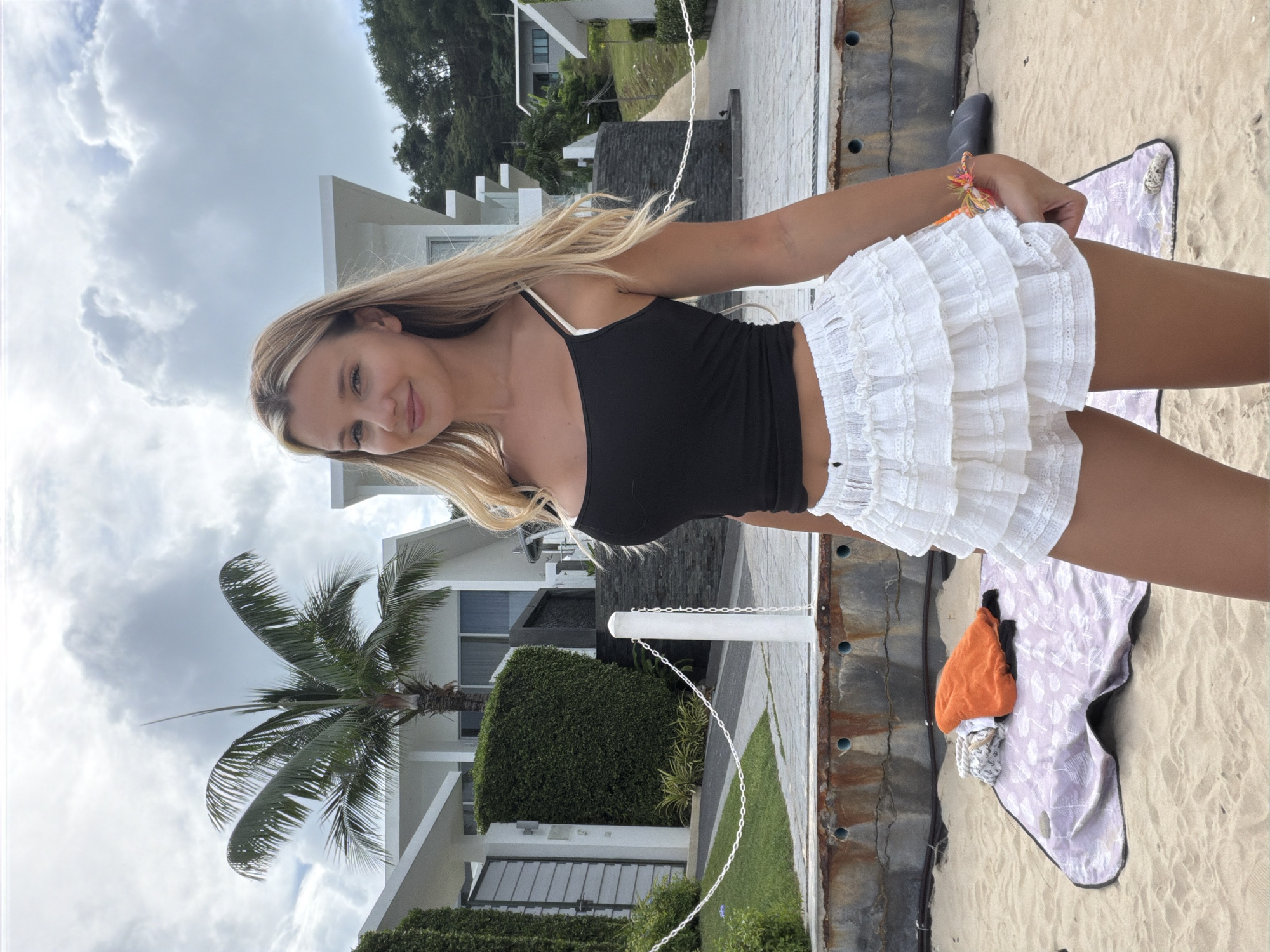 A young blonde woman stands on a sandy beach, facing the camera. She is wearing a black tank top and a white ruffled skirt. Behind her, a modern white building complex with a palm tree in front is visible. A chain link fence runs along a paved walkway in front of the building. On the sand in the foreground, a patterned blanket with some folded clothes is laid out. The sky is cloudy. This scene is located in Wichit, Thailand.