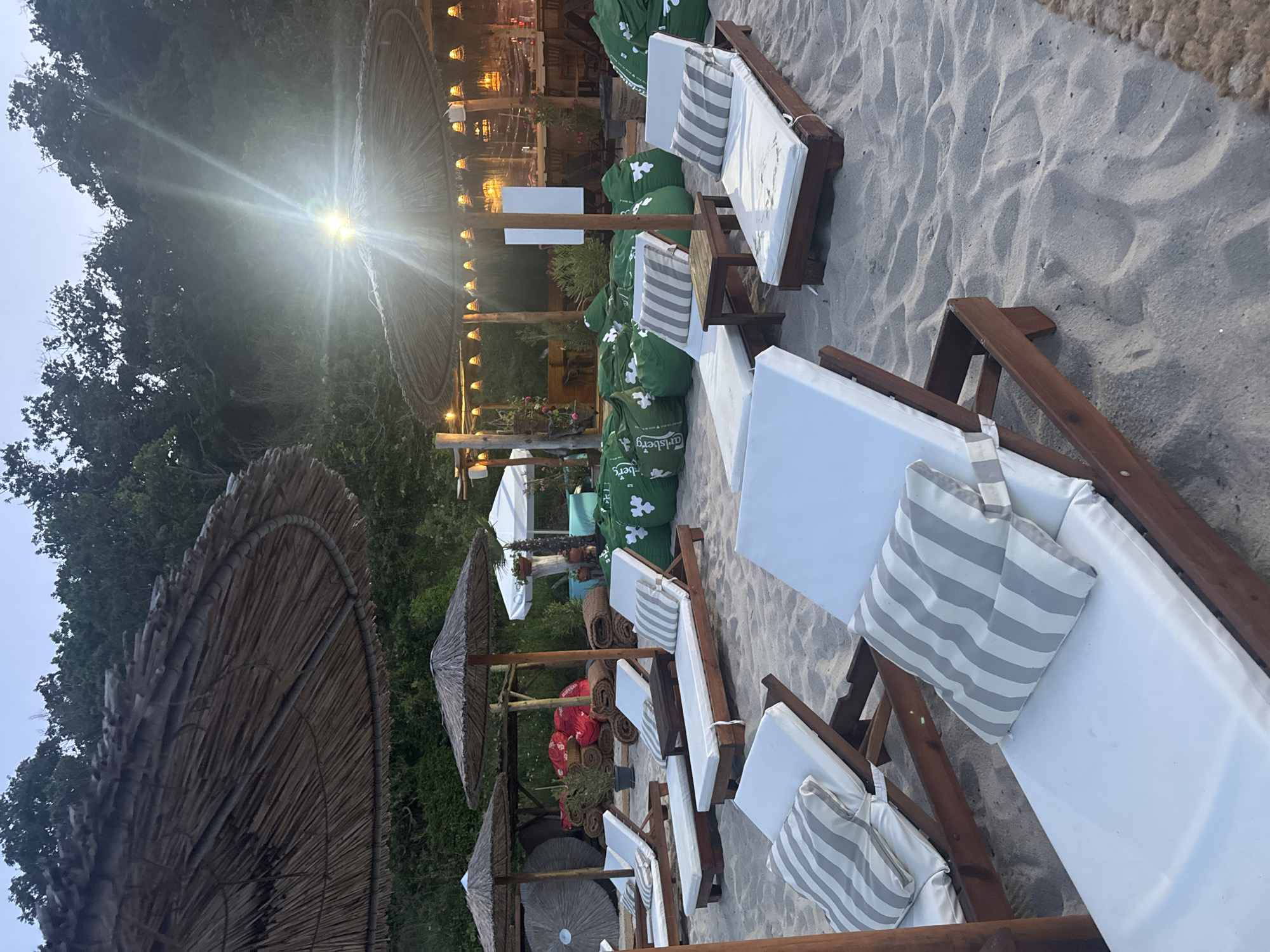 The image depicts a beach scene, likely at a resort or beach bar, featuring several sun loungers arranged on a sandy beach. In the foreground, multiple wooden sun loungers with white cushions and striped pillows are visible. Behind these, a row of large, green beanbag chairs is present, with some bearing the "Carlsberg" logo. Further back, a bar area is set up with lighting and tables. Several thatched umbrellas provide shade. The background consists of lush green trees and foliage, suggesting a natural setting. The sky appears overcast or is in the process of twilight. The overall ambiance is relaxed and inviting, typical of a seaside leisure establishment. This scene is located on the Golden Sands beach, a popular tourist destination situated north of Varna, Bulgaria.