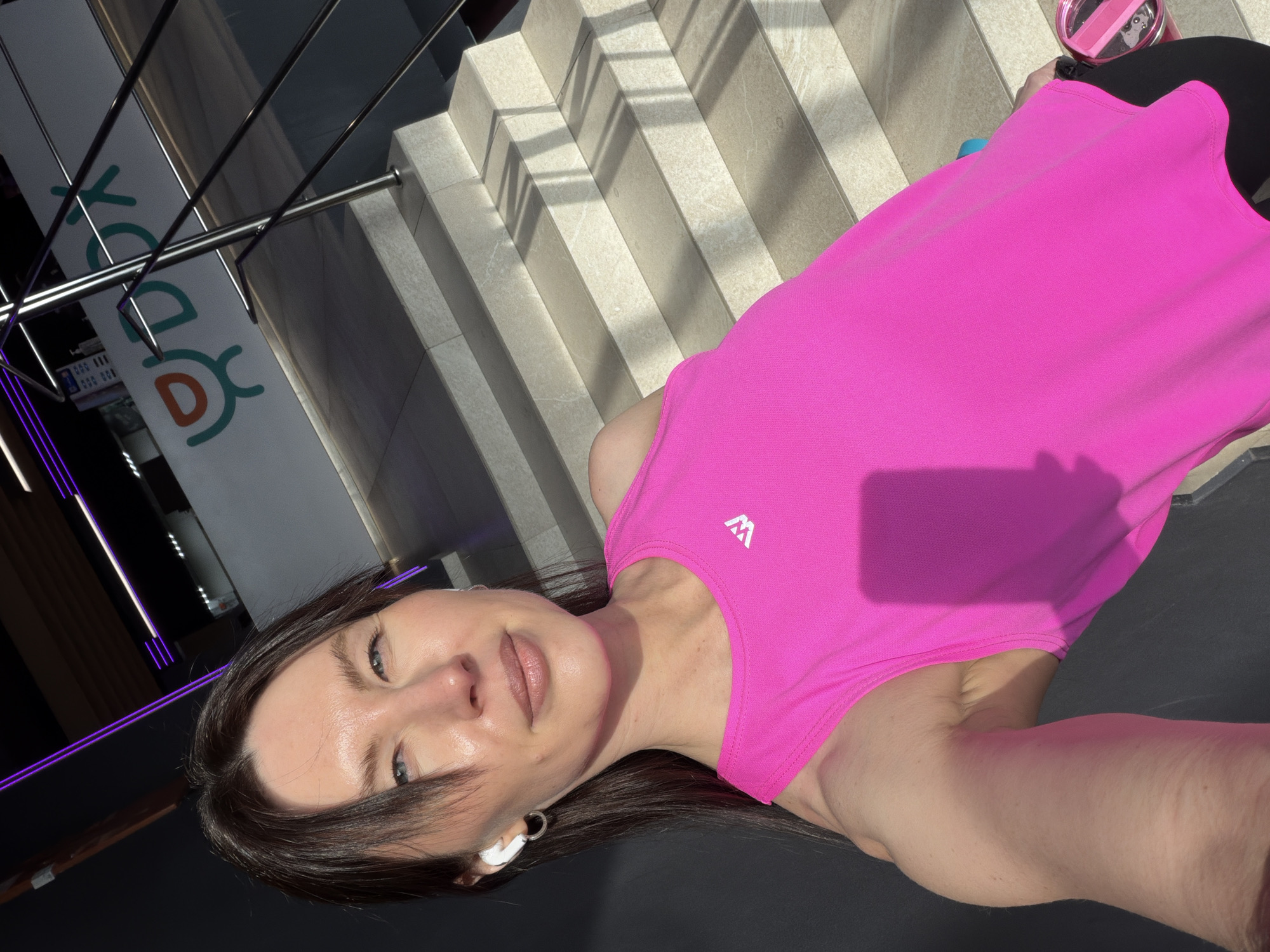 A woman with light skin, brown hair, and blue eyes is shown in a selfie. She wears a pink sleeveless top with a white logo on the left side of the chest and wireless earbuds in her left ear. Her face is slightly tilted towards the camera.
In the background are light-colored stone steps leading upwards. A branded sign is visible on the left, suggesting a commercial or public establishment. Based on the style and architecture, the image was likely taken in the city of Perm, Russia.