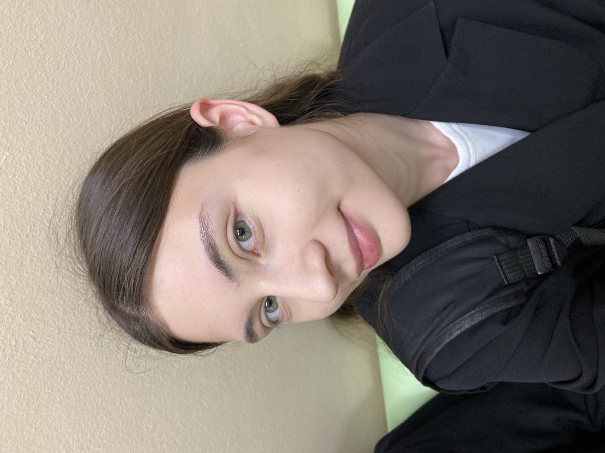 A young woman with light skin, brown hair, and green eyes is looking at the camera with a slight smile. She is wearing a black blazer and a white undershirt. Part of what appears to be a black backpack strap or bag is visible over her right shoulder. The background consists of a light yellow wall. There is no explicit indication in the image that locates the scene in Votkinsk, Russia.