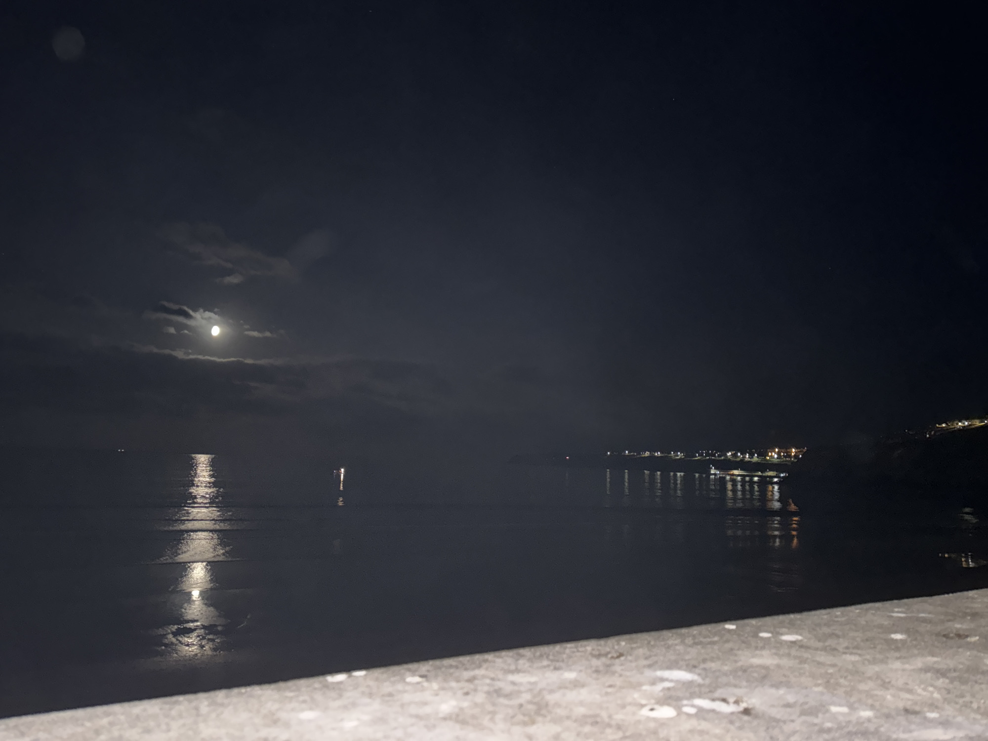 The image depicts a nighttime scene of the coast in Tramore, Ireland. The sky is dark, with a visible moon partially obscured by clouds, casting a luminous reflection on the calm water. The water's surface is also illuminated by distant lights from a town or village situated along the coastline. These lights create shimmering reflections that stretch across the water. In the foreground, a textured concrete surface is visible, suggesting a pier, promenade, or a similar man-made structure overlooking the sea. There are no people or specific animals identifiable in the image. The overall atmosphere is serene and tranquil, characteristic of a quiet night by the sea.