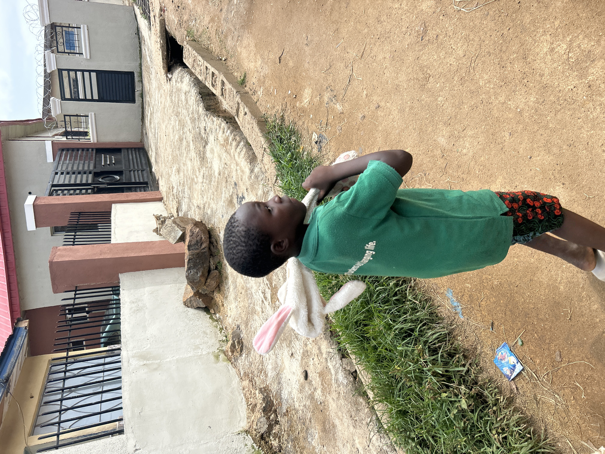 A young boy, likely of Nigerian descent, walks on a dirt path in Jos, Nigeria, carrying a white plush toy rabbit with pink ears over his right shoulder. He is wearing a green short-sleeved polo shirt and dark shorts with a red and black floral pattern. The boy is looking to his right, away from the camera. In the background, there is a beige and red building with barred windows and a metal gate. A drainage ditch runs along the right side of the path. The ground is a mix of dirt and concrete. The sky is overcast.
