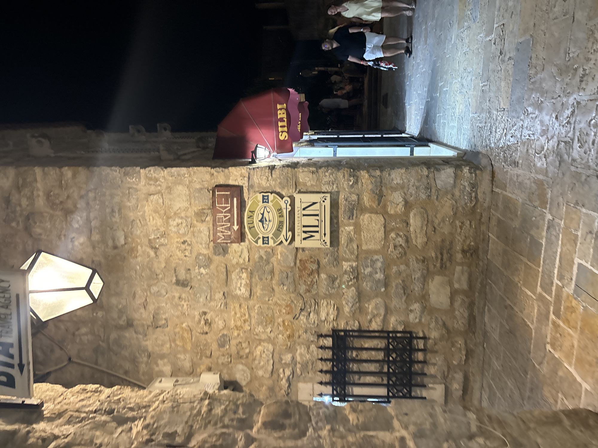 The image depicts a narrow alleyway in Budva, Montenegro, at night. A stone wall dominates the left side of the frame, adorned with several signs: "Market," a circular sign featuring a fish, and "MLIN". A lantern is mounted on the wall. Above the signs, a red awning marked "SILBI" protrudes into the alley. Several people are standing at the end of the alleyway. The cobblestone street appears slightly wet. The overall lighting is dim.