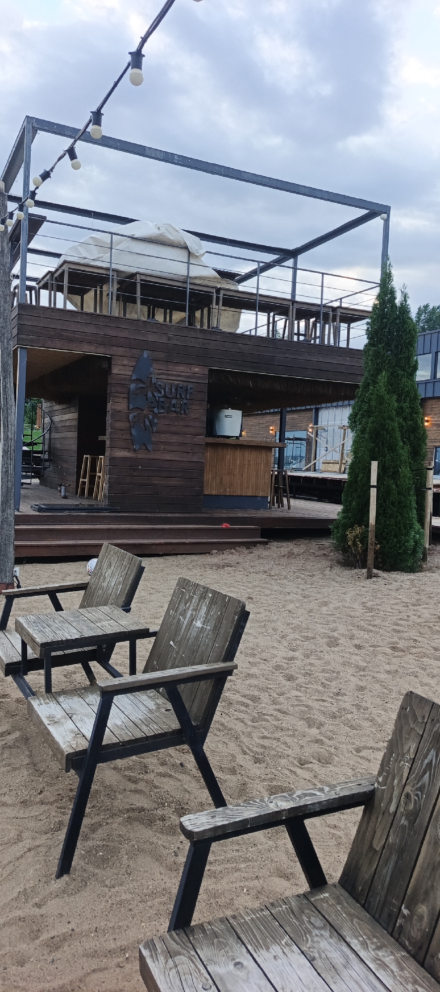 The image shows an outdoor seating area with wooden chairs and tables placed on a sandy ground. In the background, there is a two-story wooden building with a metal frame structure. The building has signage that reads "SURF BAR". There are also trees and outdoor lighting visible. The sky is overcast. The scene appears to be a recreational area, likely a bar or cafe, by a body of water, possibly a beach, given the sandy ground and the presence of chairs and tables for outdoor seating. The location is identified as Votkinsk, Russia.