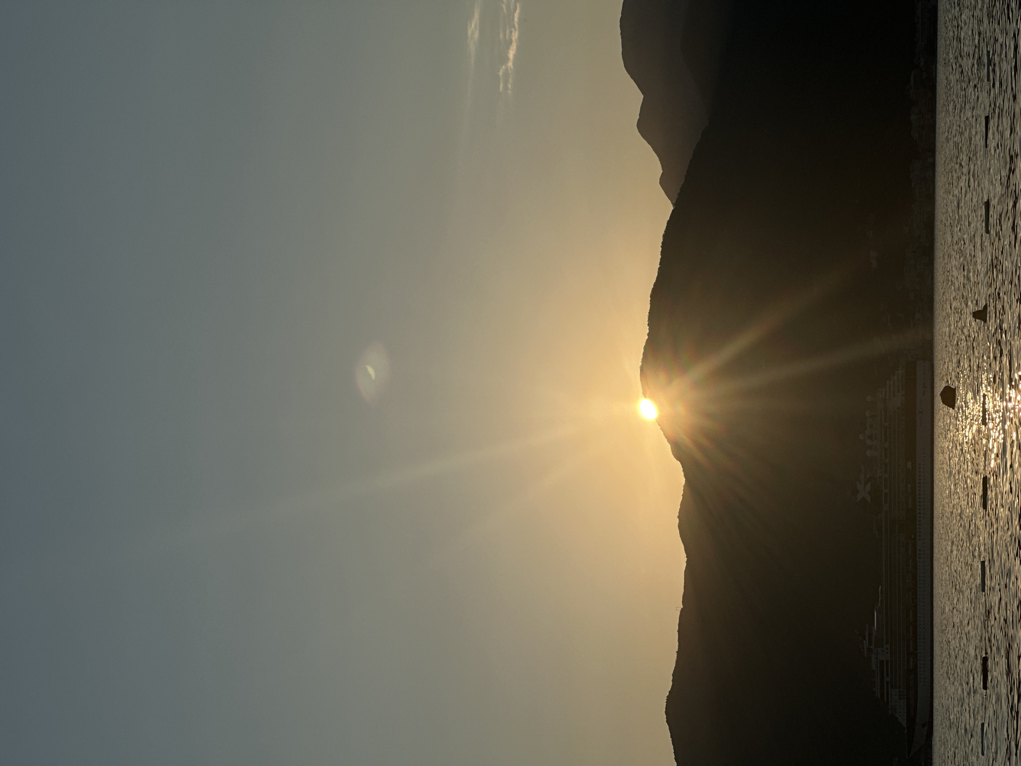 This is a landscape photograph captured during sunset in Donja Lastva, Montenegro. The scene depicts a dramatic silhouette of a mountain range on the right side of the frame, with the sun setting behind its peaks. The low angle of the sun creates strong sunbursts and lens flares that illuminate the sky with warm, golden hues.
In the foreground, a calm body of water reflects the golden light, creating a shimmering effect. Several buoys are visible, marking a swimming or boating area. A large cruise ship is docked or anchored along the coastline, partially obscured by the mountain's shadow and the low sunlight. Some structures, possibly buildings or piers, are visible along the shore at the base of the mountain. The sky is a gradient of soft blues and yellows, with a few wispy clouds. The overall mood is serene and picturesque, characteristic of a peaceful evening by the sea. The time of day is clearly sunset.