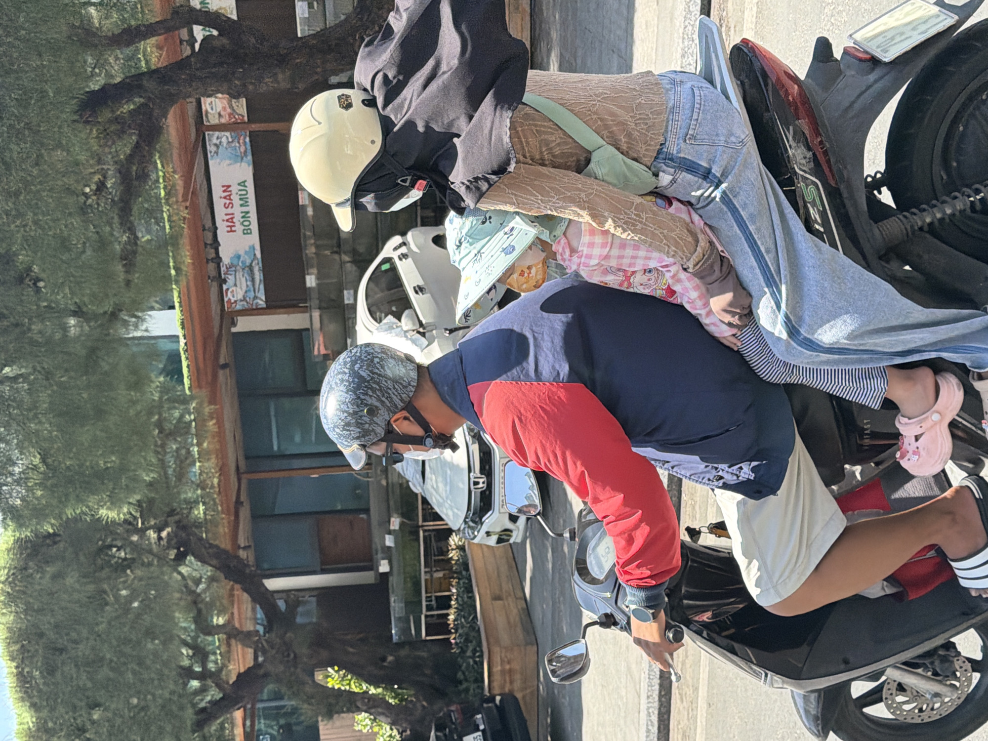 On a sunny day in Nha Trang, Vietnam, three people are riding on a black scooter. The rider, a man, wears a camouflage helmet and a red and blue jacket. He is looking forward. Behind him sits a woman, wearing a light-colored helmet and a beige long-sleeved shirt with a floral pattern. She is holding a child in front of her. The child is wearing a pink and white checkered shirt and a light blue hat. The scooter is on a paved road with trees and buildings in the background. A white car is parked behind the scooter. The area appears to be a commercial district, possibly near a seafood market as indicated by a sign in Vietnamese.