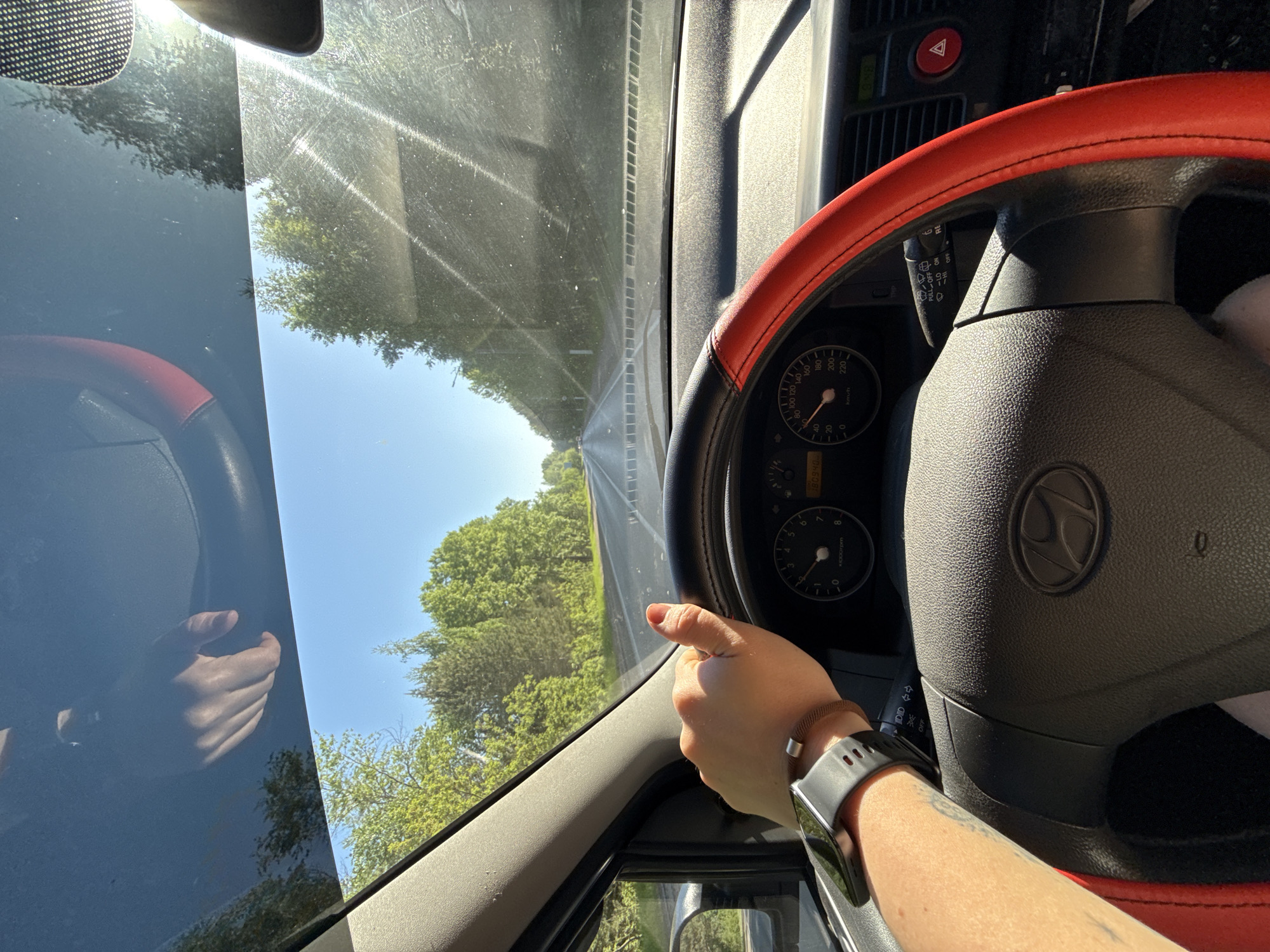 A driver's point-of-view photo taken inside a car on a road bordered by trees, presumably in or near the city of Chaikovsky, Russia. The driver's hand is visible on the right side of the steering wheel, adorned with a bracelet and a smart watch. The car's dashboard and speedometer are visible. The sky is blue and sunny.
