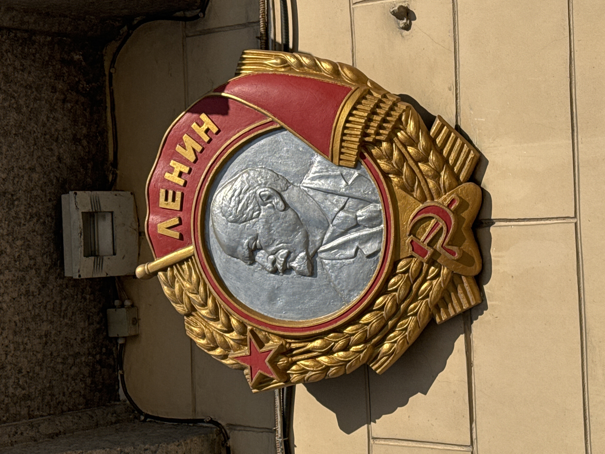 A Soviet-era monument honoring Lenin is affixed to a building in Saint Petersburg, Russia. The monument is circular, with a raised image of Lenin's head in profile at the center, surrounded by a wreath of golden wheat. Above the portrait is a red banner with the name "Ленин" (Lenin) in gold Cyrillic letters. A red star is visible at the bottom, and a hammer and sickle are at the upper right. The monument is mounted on a beige-colored wall with vertical paneling. To the left of the monument are various electrical fixtures and conduit attached to a textured, darker-colored stone wall. The scene is brightly lit, casting shadows from the monument onto the wall.
