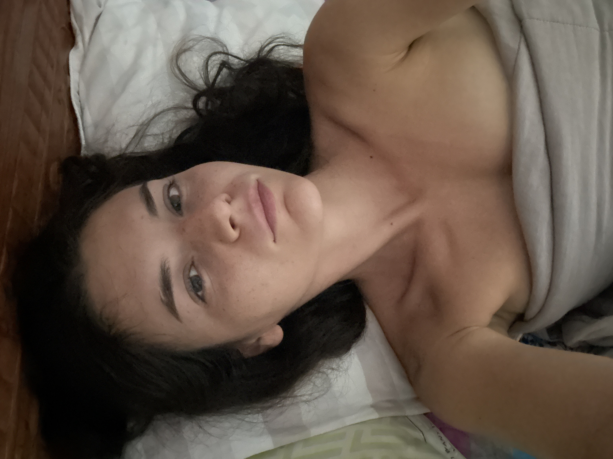 A woman with dark hair and freckles is lying on a bed with a white and green striped sheet and a white pillow. She is partially covered with a gray blanket. The location cannot be determined from the image.