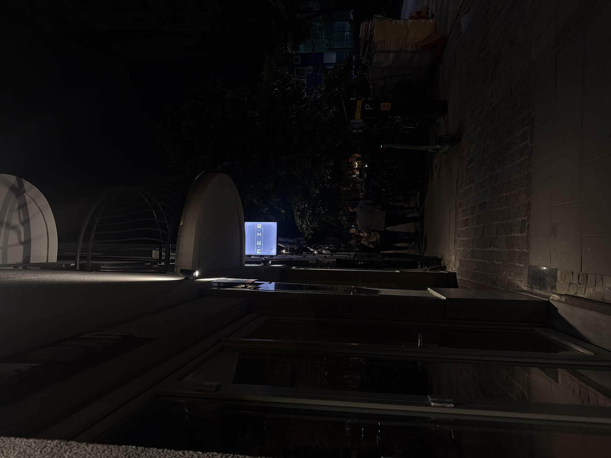 Captured at night in an urban setting, the image presents a low-angle view towards a building with rounded architectural features on the left. A sign, brightly lit with the word "MEZIE" visible, stands prominently in the middle of the frame. Beyond the sign, the background reveals foliage and urban elements, possibly other buildings. The foreground is dominated by what appears to be a window or another architectural element. A group of people is standing on the right.