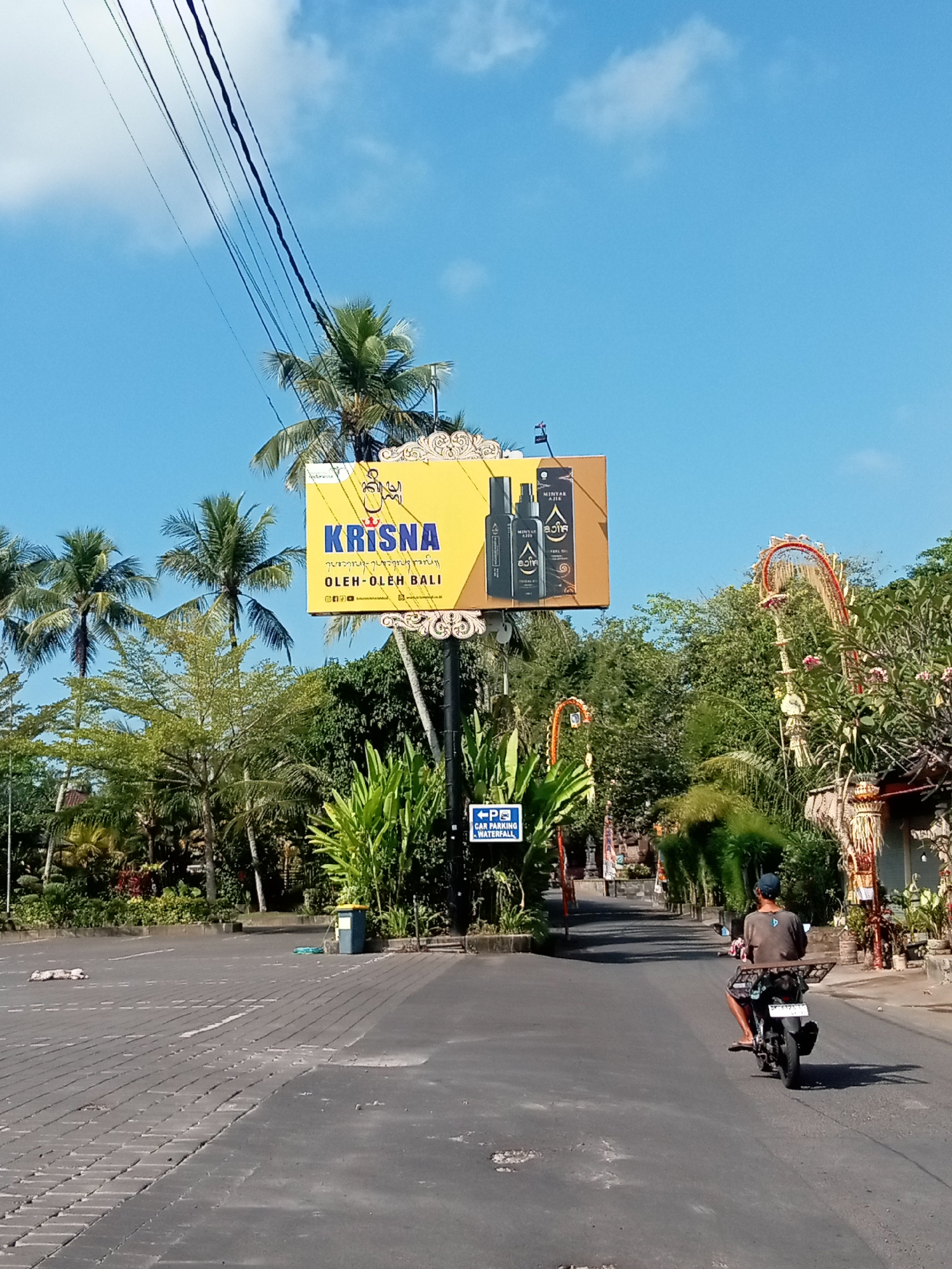 A bright yellow billboard displays "KRISNA OLEH-OLEH BALI" with product images and is situated against a clear blue sky, partially obscured by palm trees and electrical wires. Below the billboard, a street curves towards the background, lined with lush green foliage and ornate decorative structures. A person on a motorcycle, wearing a dark t-shirt and a blue cap, rides away from the viewer down the street. A blue sign indicating "CAR PARKING WATERFALL" is visible on the left side of the road. The foreground shows a paved road with a brick-like pattern.