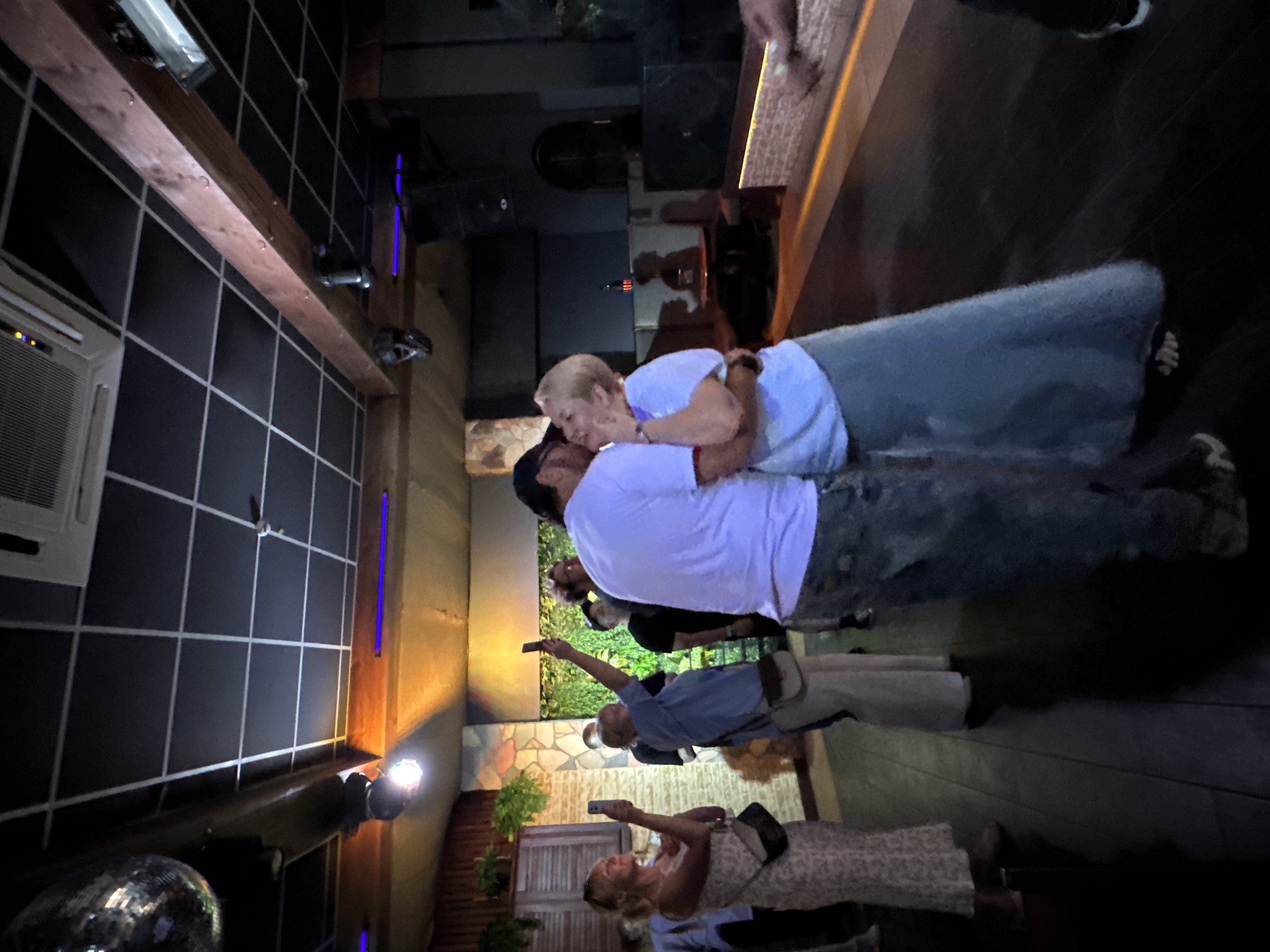 The image shows a crowded bar interior in Chaikovsky, Russia. Several patrons are present, taking photos with their smartphones. A man and an older woman are embracing near the bar. The lighting is dim, with various light fixtures illuminating the scene. The walls feature different textures and materials, including tiles and stone. A disco ball is partially visible in the bottom left corner.