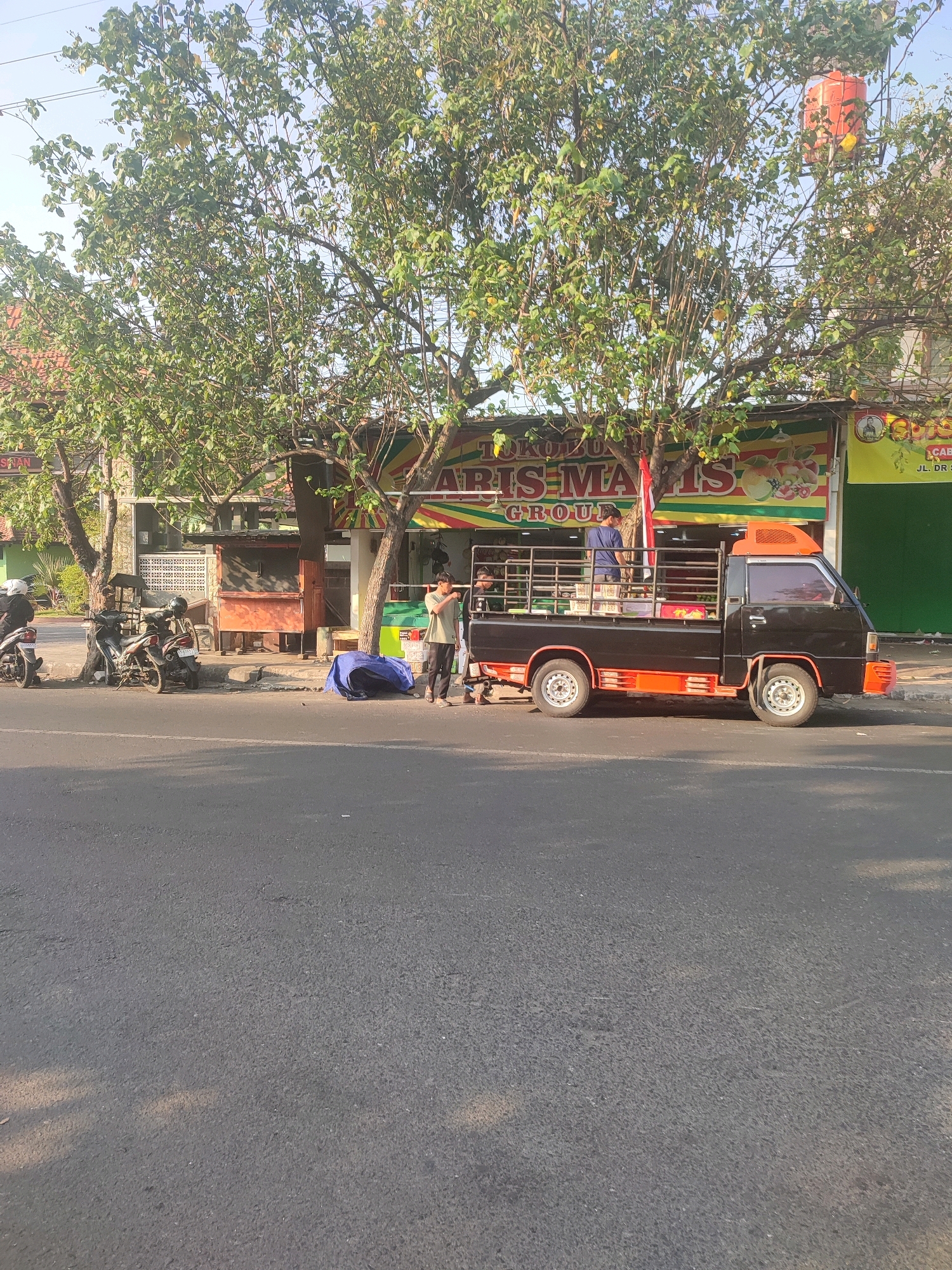 In the city of Nganjuk, Indonesia, an outdoor scene unfolds along a paved road. Sunlight filters through the leaves of a large tree, casting dappled shadows on the asphalt. To the right, a black pickup truck with orange accents is parked at the roadside. A man in a light green t-shirt and dark pants stands beside the truck, appearing to be engaged in conversation. He is holding a phone in his left hand. Behind him, another person in a blue shirt is partially visible in the truck bed, which is equipped with a metal cage.
In front of the truck, a blue tarp is spread on the ground, and a man in a blue shirt is seen standing next to it. To the left of the truck, two motorcycles are parked on the sidewalk. One is black with a rider wearing a white helmet, and the other is also black. A building with a sign reading "ARIS MAJIS GROUP" is visible behind the truck, featuring colorful imagery and text in Indonesian. Adjacent to this building, a green structure and an orange stall are also present. The overall impression is a street scene in a tropical environment, likely during the daytime.