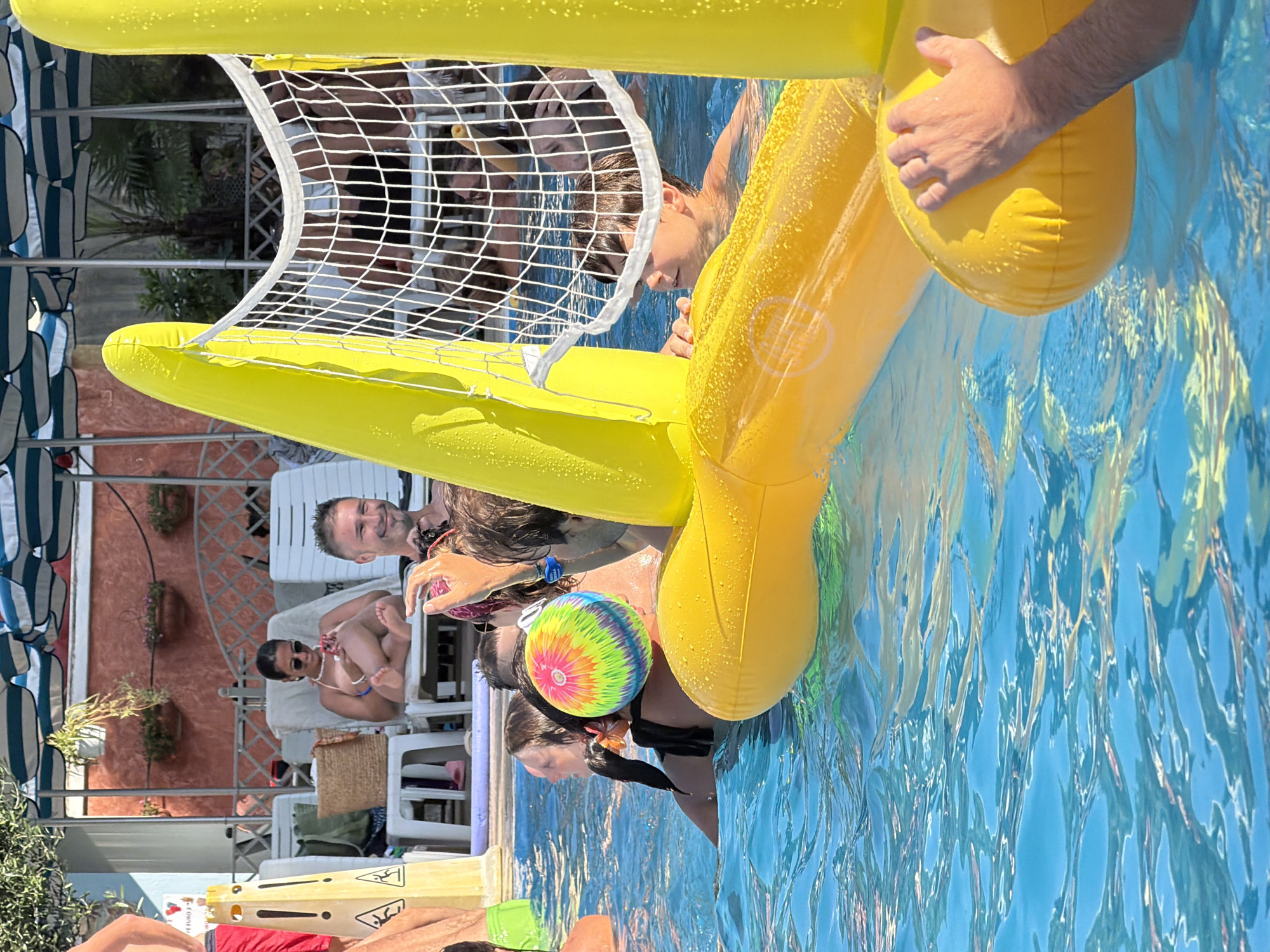 This image depicts an outdoor swimming pool scene at a location in Gatteo, Italy, likely a resort or hotel. In the foreground, an inflatable yellow volleyball net structure is partially submerged in the bright blue water. Several people are in the pool, some partially visible. To the left, a person wearing green shorts is in the water. In the center, a person with dark hair is seen from the back, wearing a black bikini top, with a colorful tie-dye ball behind their head. To the right, a man's hand is visible resting on the inflatable structure. In the background, a woman is seated on a lounge chair, looking at her phone, while other lounge chairs and umbrellas are visible, suggesting a recreational area. The overall atmosphere is relaxed and summery.