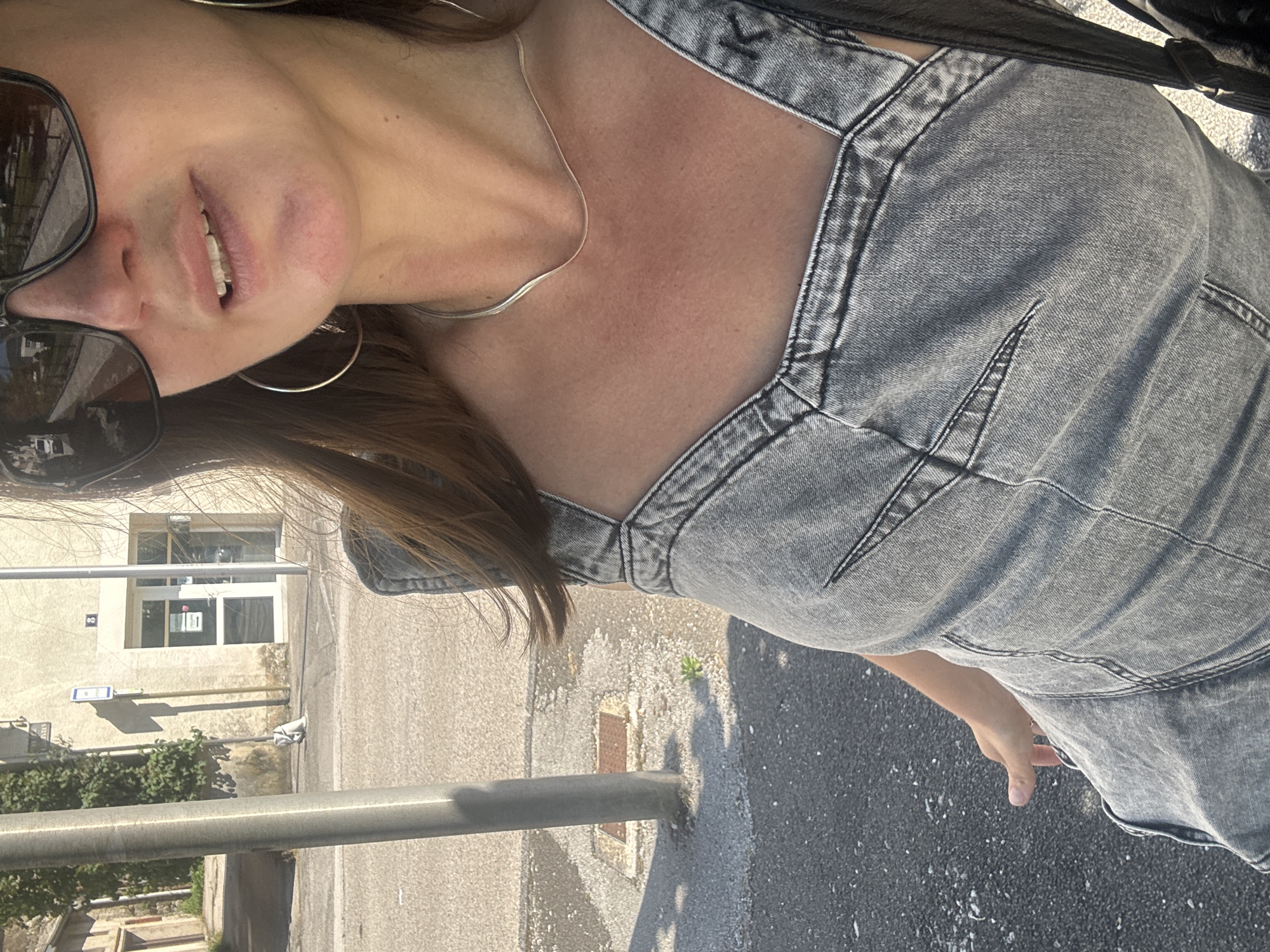 A woman wearing large sunglasses, hoop earrings, a silver necklace, and a grey denim dress stands outdoors on a sunny day. She is looking to her right, with her mouth slightly open. The background shows a building with a blue sign and windows, a street, and a lamppost. This scene appears to be in the city of Saint-Claude, France, given the architectural style and the presence of a building number "3" visible on the facade.